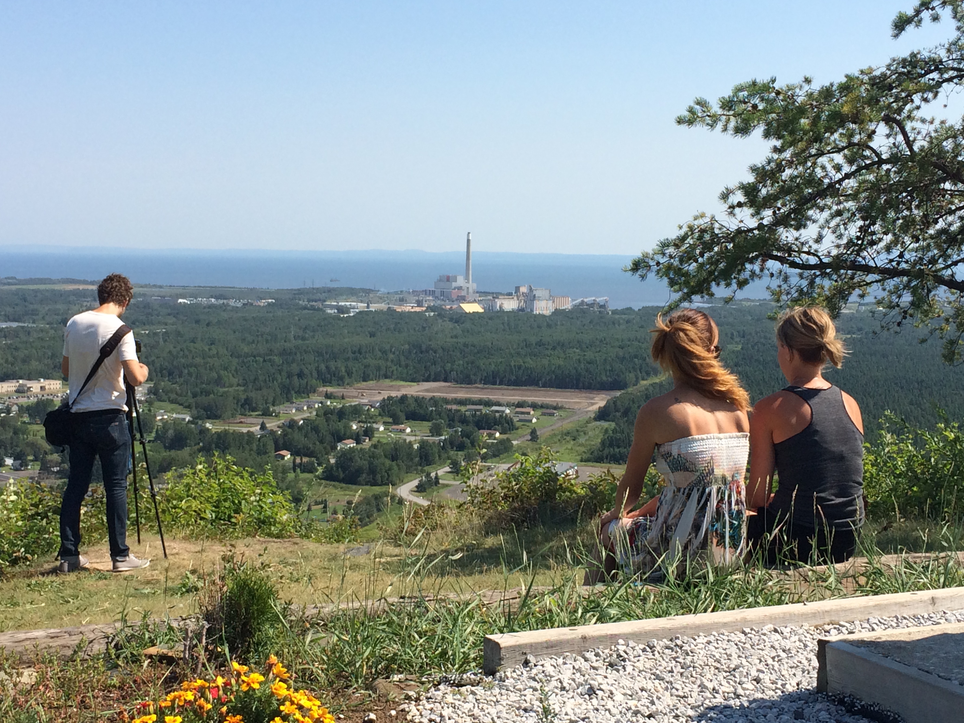 two young women sit in the sun on a grassy lookout overlooking Thunder Bay. A young man stands at the edge of the lookout, taking a photograph of the view. 