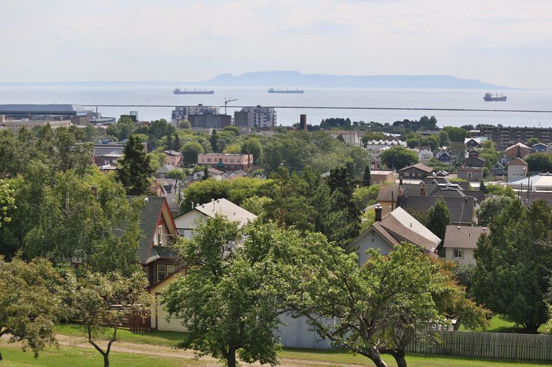 an aerial shot of the city of Thunder Bay; a cityscape with houses and greenery progressing to a harbour on Lake Superior in the distance. The faint misty outline of Sleeping Giant is on the horizon.