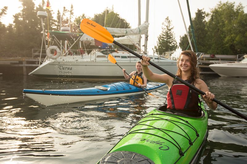 two smiling people in kayaks paddle in a harbour in golden late afternoon light on a summer day.