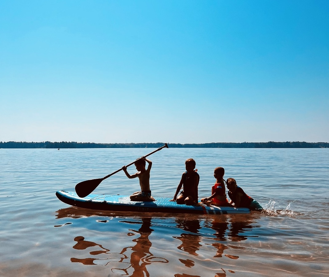 3 children and a dog sitting on a paddleboard in a blue lake under a clear blue sky. The child in front is holding up a large paddle. 