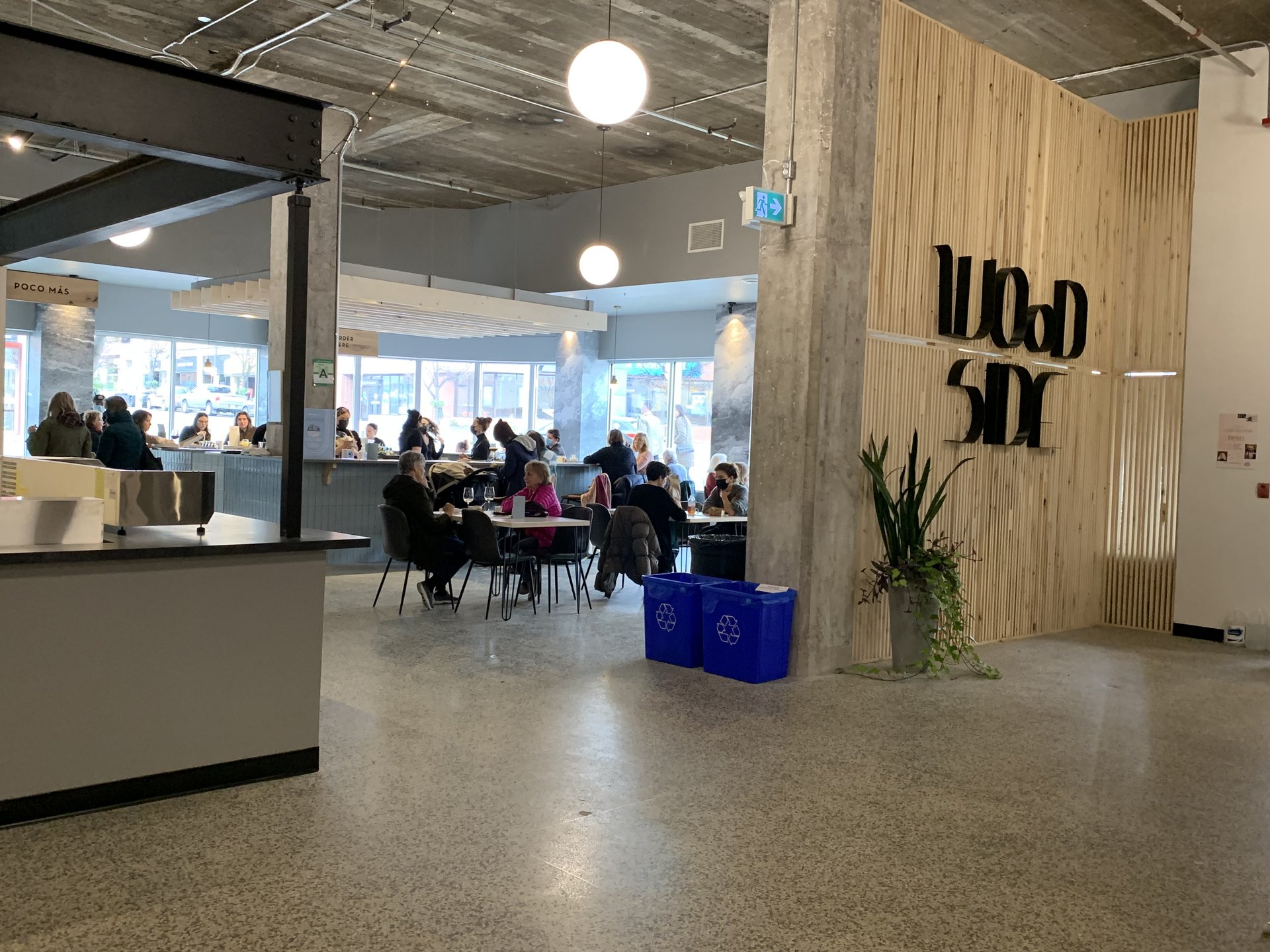 Woodside at Goods & Co; a wooden slat wall with a sign that reads "Woodside" that opens into a large, well-lit meeting space filled with tables, chairs and people. 