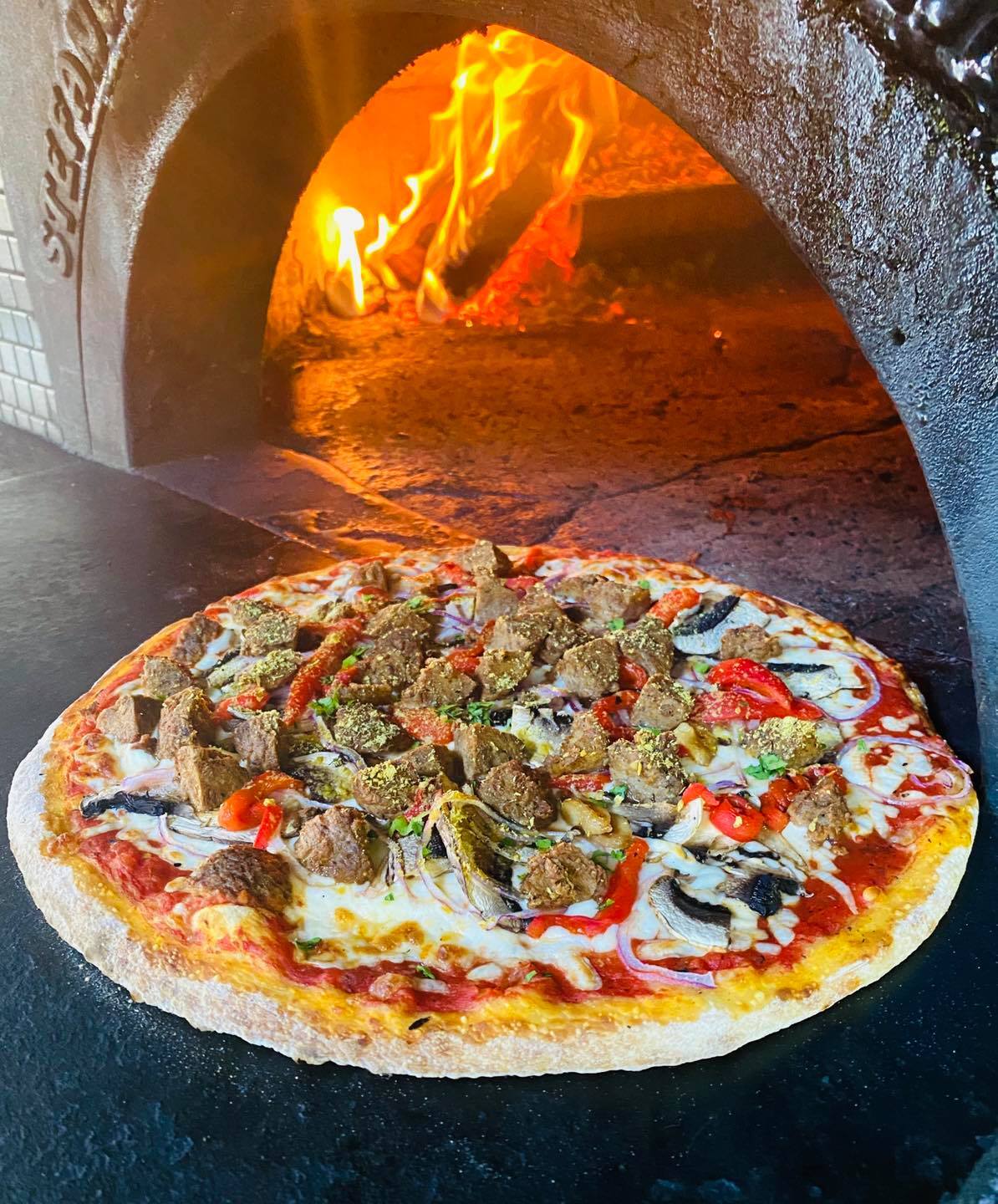 a vegan pizza covered in fresh, colourful toppings next to a glowing hot wood-fired clay oven.