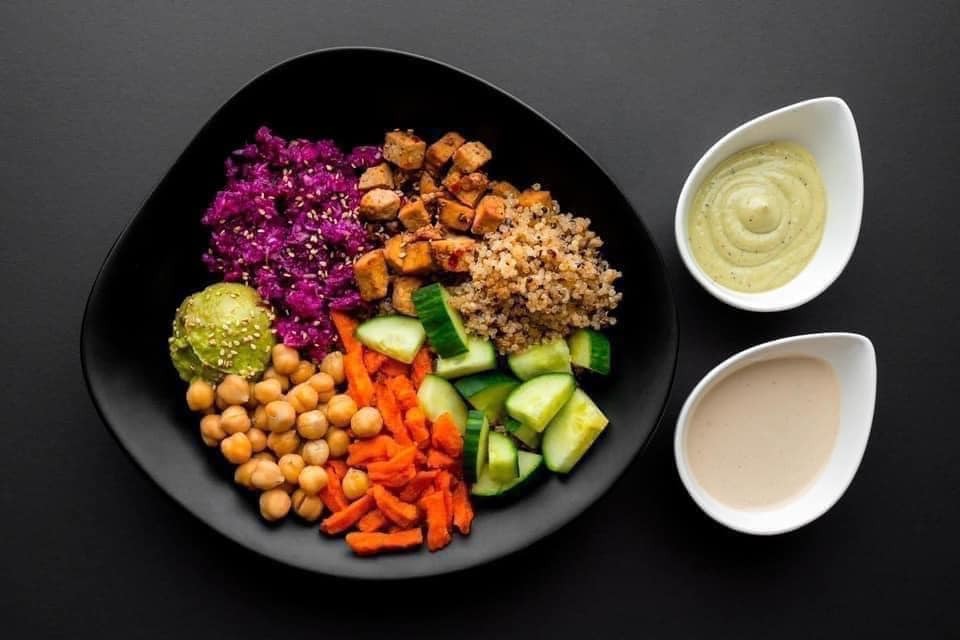 a Buddha Bowl from gd2go restaurant; a black bowl on a black background filled with quinoa, chick peas, diced carrots, cucumber and more, with two delicate white egg-shaped ramikens of sauces on the side. 