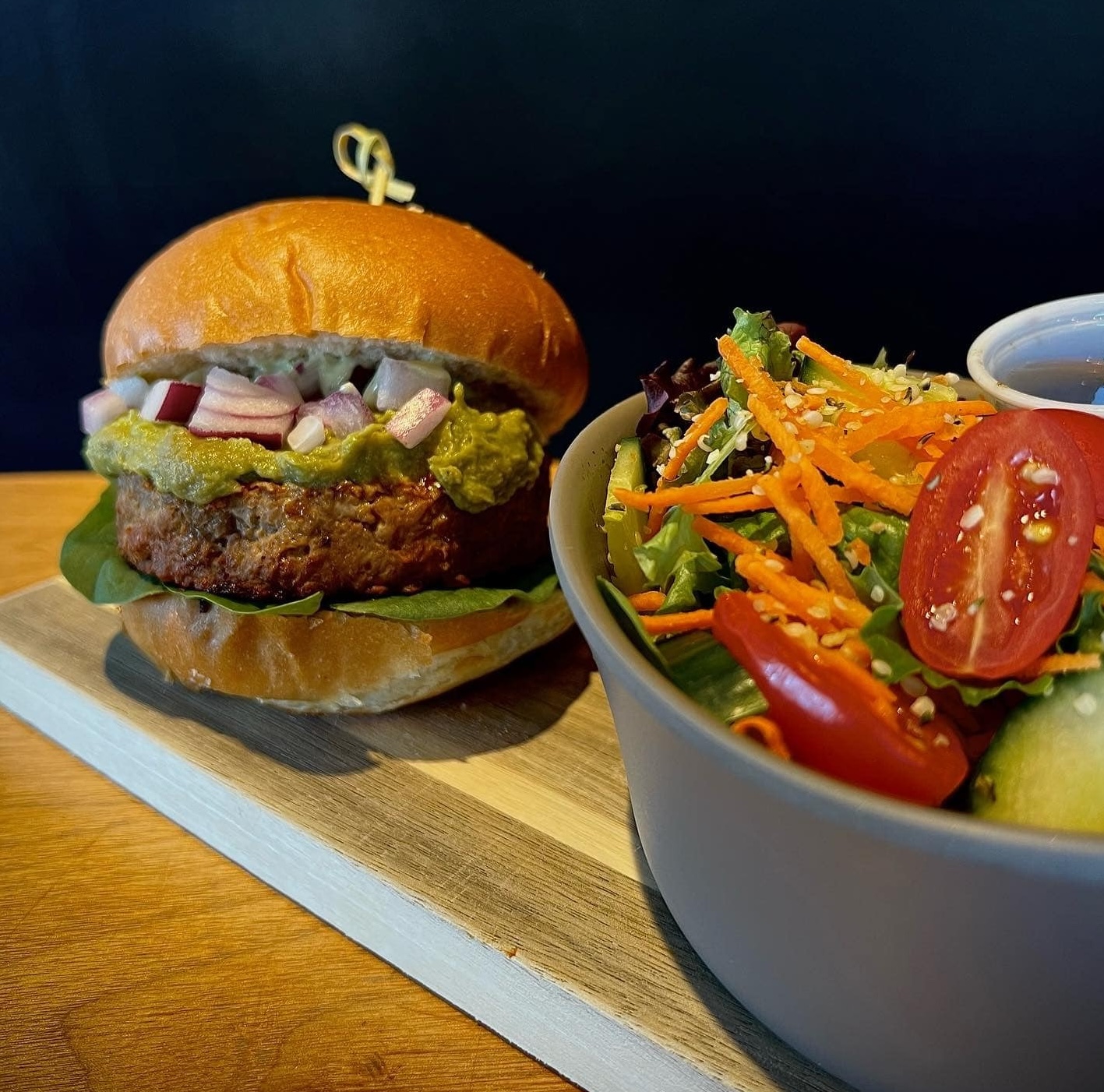 a vegan burger stacked with guacamole and fresh vegetables next to a large, crisp and colourful salad.