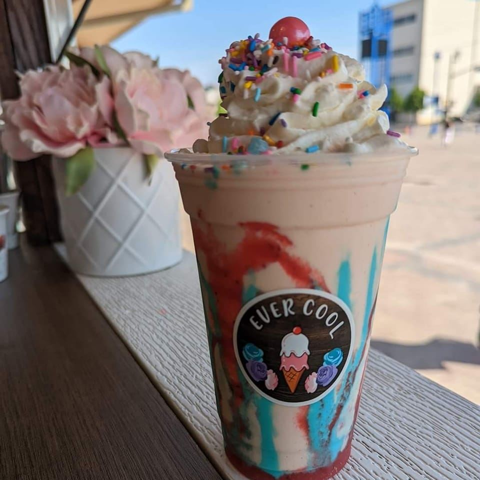 a colourful milkshake topped with whipped cream, sprinkles and a cherry, sitting next to a vase of pink flowers on a windowsill. A bright blue sky and sunshine is in the background.