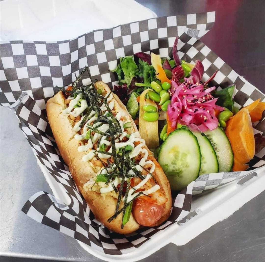 a plant-based vegan hot dog with kimchi and sliced veggies on the side on checkered wax paper in a to-go container.