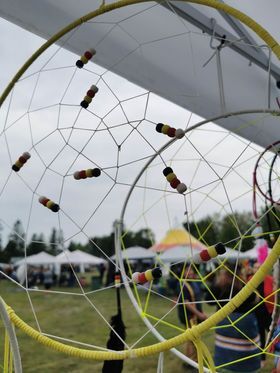 a closeup of two dreamcatchers hanging in an outdoor vendor booth, with a ring of vendor tents, green grass and trees in the background.