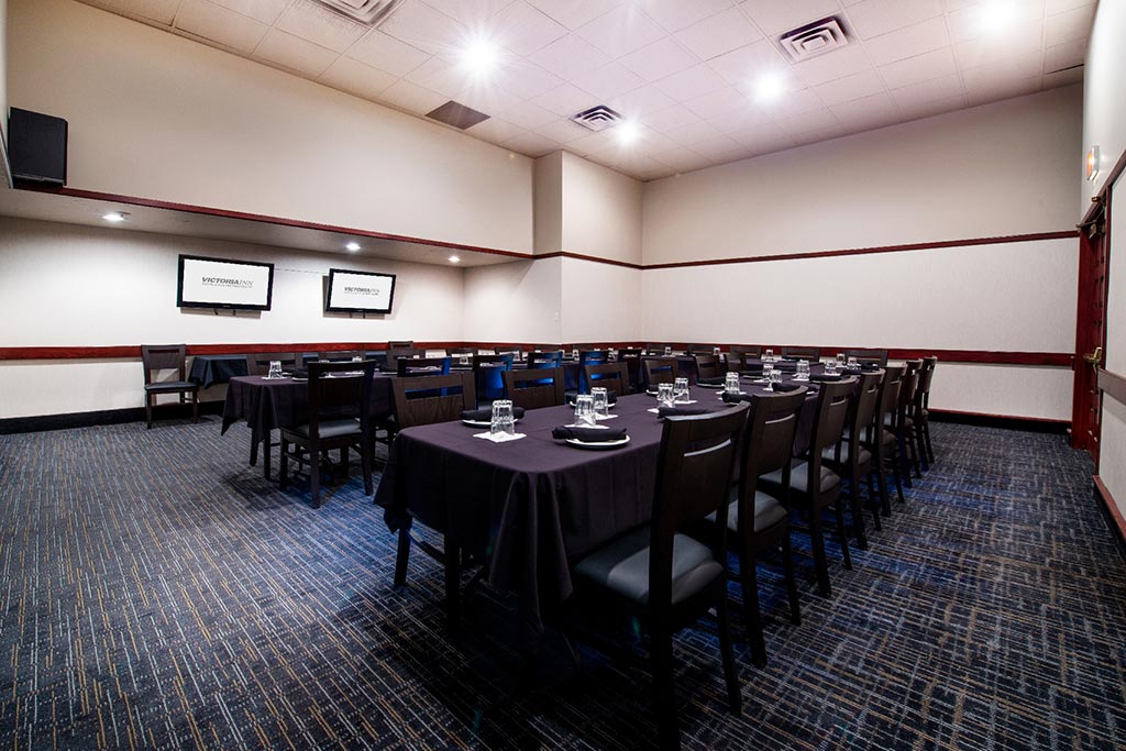 A meeting room at the Victoria Inn Thunder Bay; a large, professional-looking meeting room with long tables adorned with dark purple table cloths and glasses, boardered by chairs. It is well lit and has a dark patterned carpet.