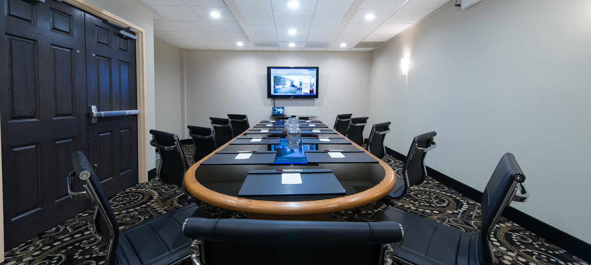 The boardroom at the Victoria Inn Thunder Bay; a polished and modern-looking meeting room with long glass and wood table surrounded by black chairs, a black patterned carpet and brightly lit by pot lights.  