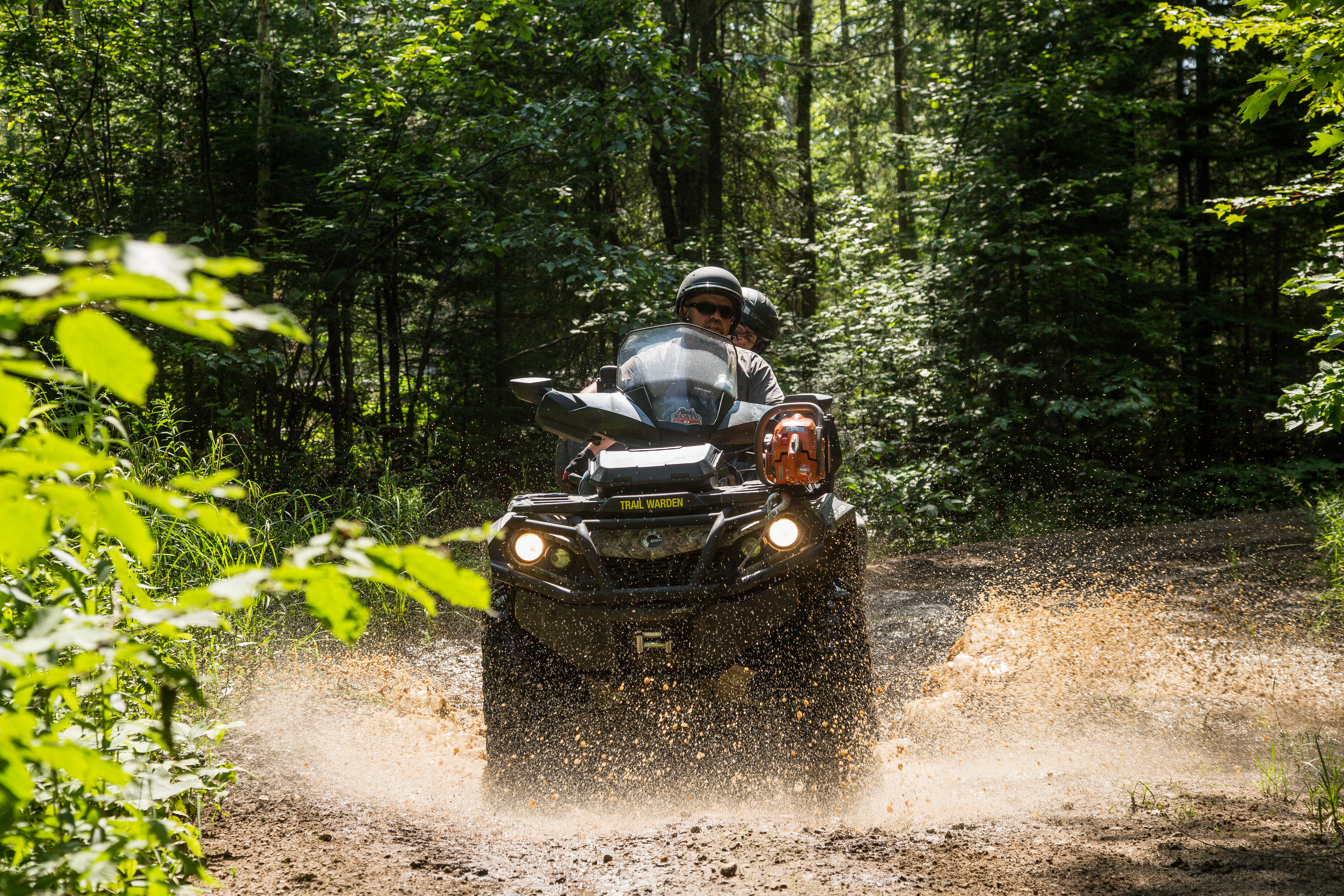An ATV with two riders rolling down a dirt trail on a sunny day, surrounded in tall, lush green forest. The ATV is splashing up muddy water that catches the sunlight and appears to glow.