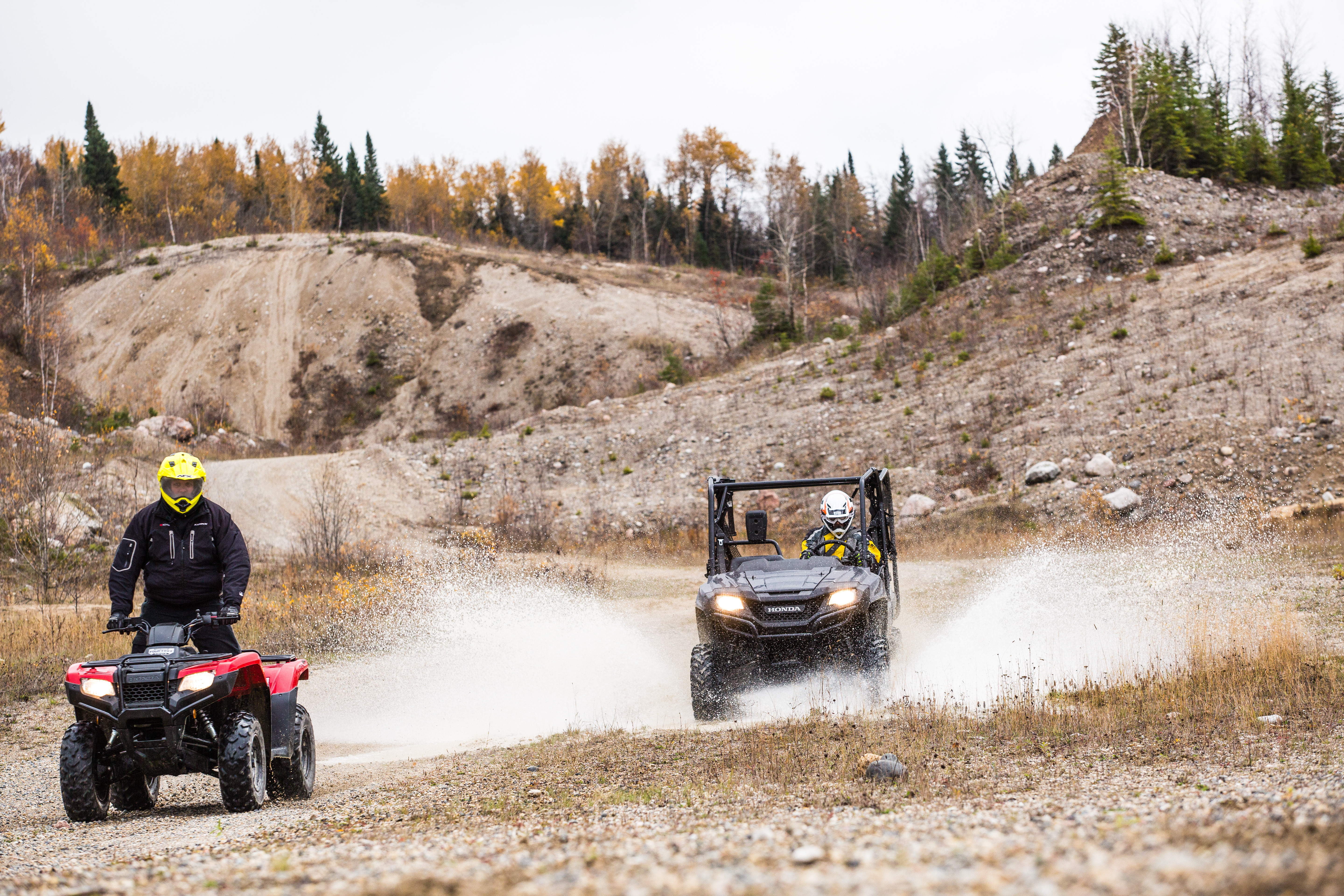 An ATV and a UTV drive through a series of gravel hills, kicking up clouds of dust as they go. 