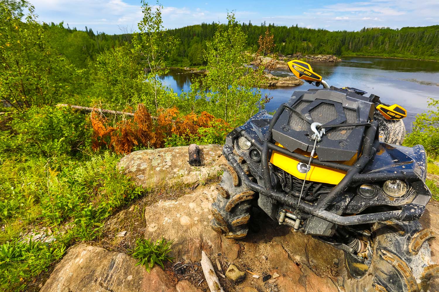 A muddy ATV with a winch on the front parked on an uneven, rocky bank covered in weeds and brush. In the background there is a lake surrounded by green forest.