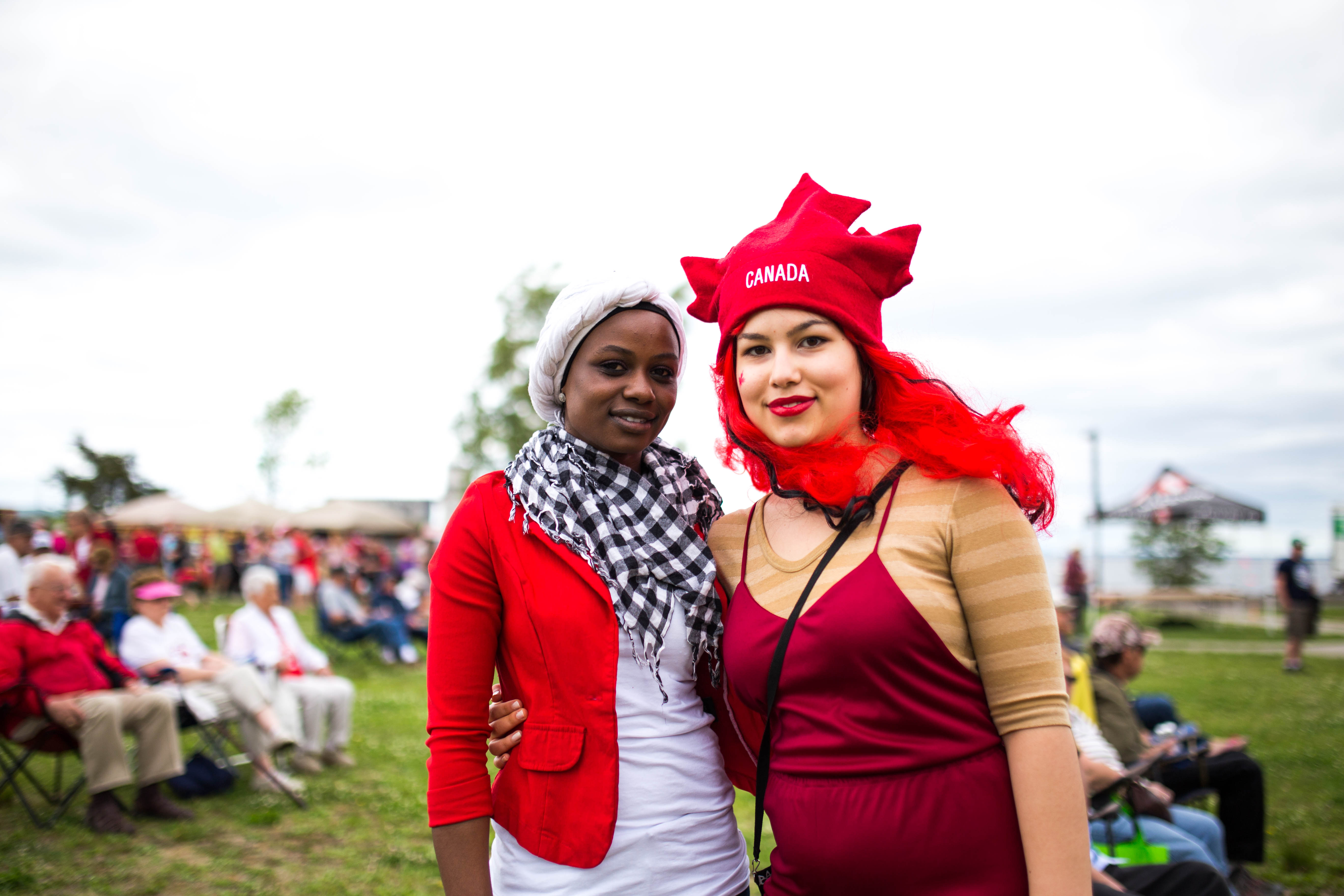 Two young women wearing red and white and one wearing a hat shaped like a maple leaf, smiling with their arms around each other's shoulder. 