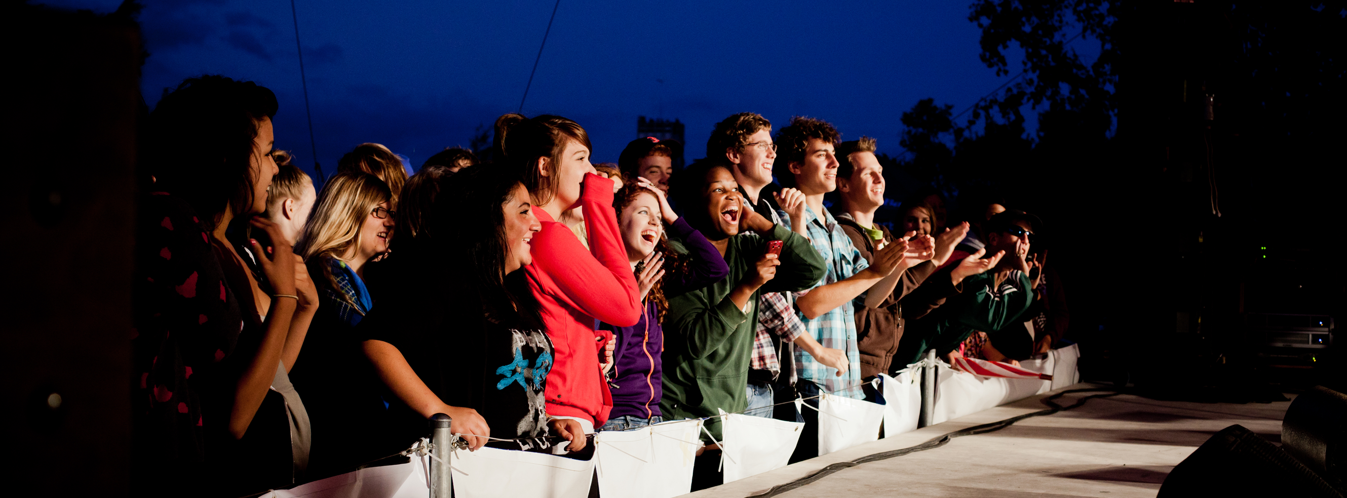 A cheering, smiling crowd lined up in front of an outdoor stage at night.