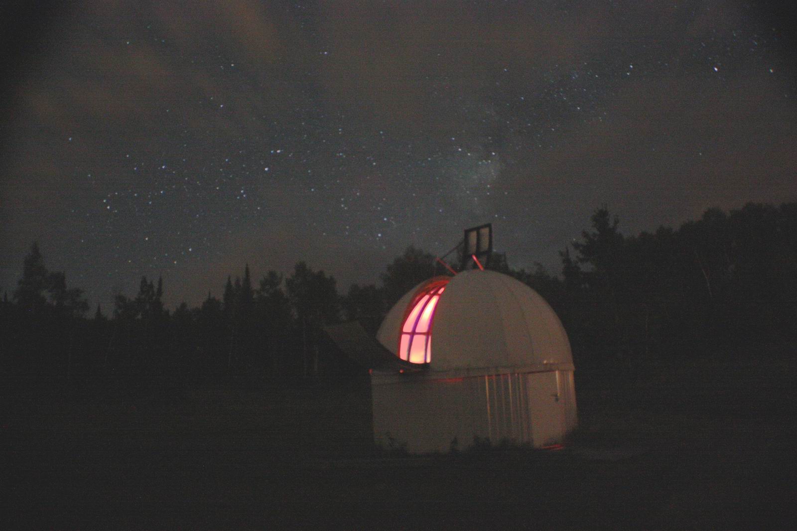 The David Thompson Observatory at night; a white metal dome-shaped building with a lit window at the top which exposes the telescope. There is a mist of stars in the night sky overhead and the dark silhouette of forest in the background.