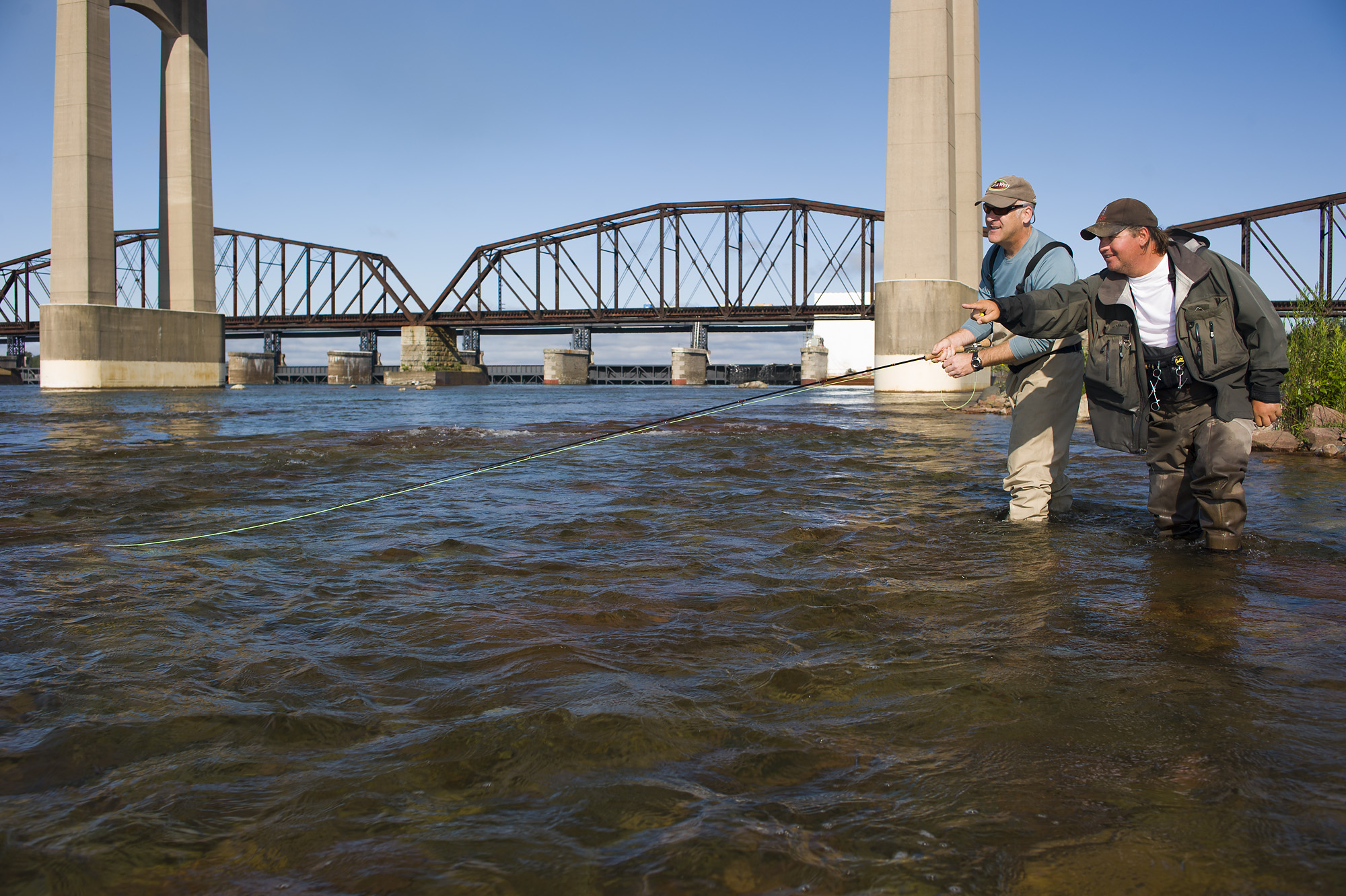 two men fishing in St. Mary's River next to the footings of a large bridge in Sault Ste. Marie. Another bridge and a clear blue sky are in the background.