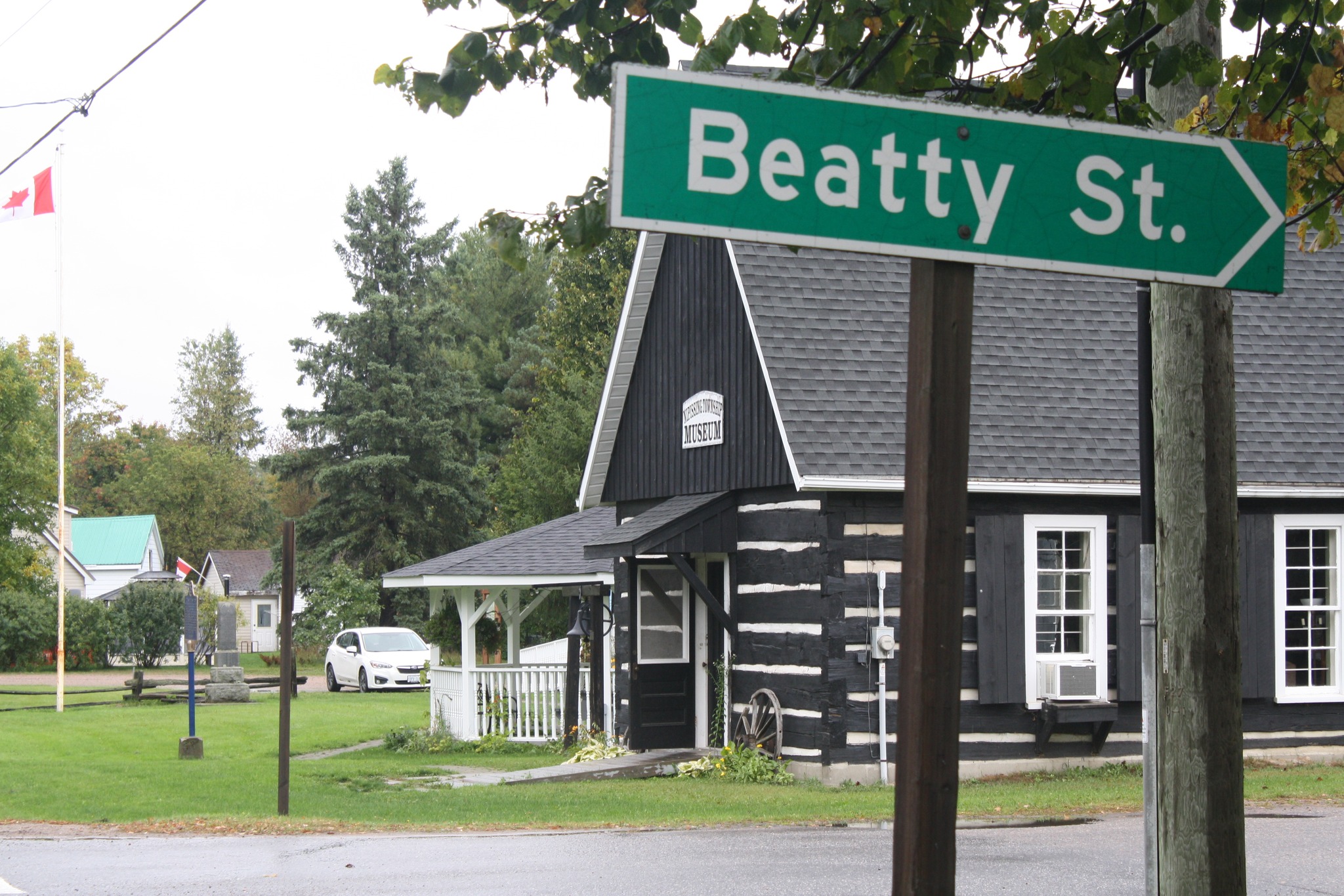 a brown and white painted wooden log A-frame church from the early 1900s, now part of the Nipissing Township Museum. A street sign in the foreground reads "Beatty St.".