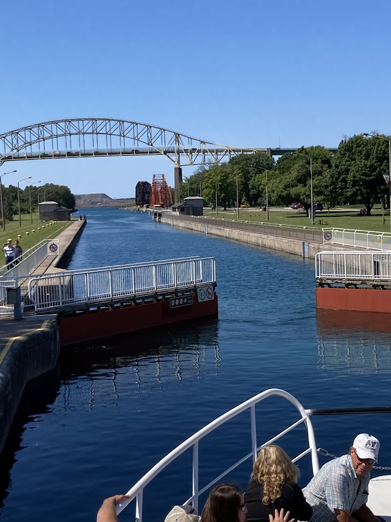 the Sault Locks opening to let a ship through, as passengers on the ship stand at the railing to watch. It is a sunny summer day and the water in the lock is reflecting the very blue sky. 