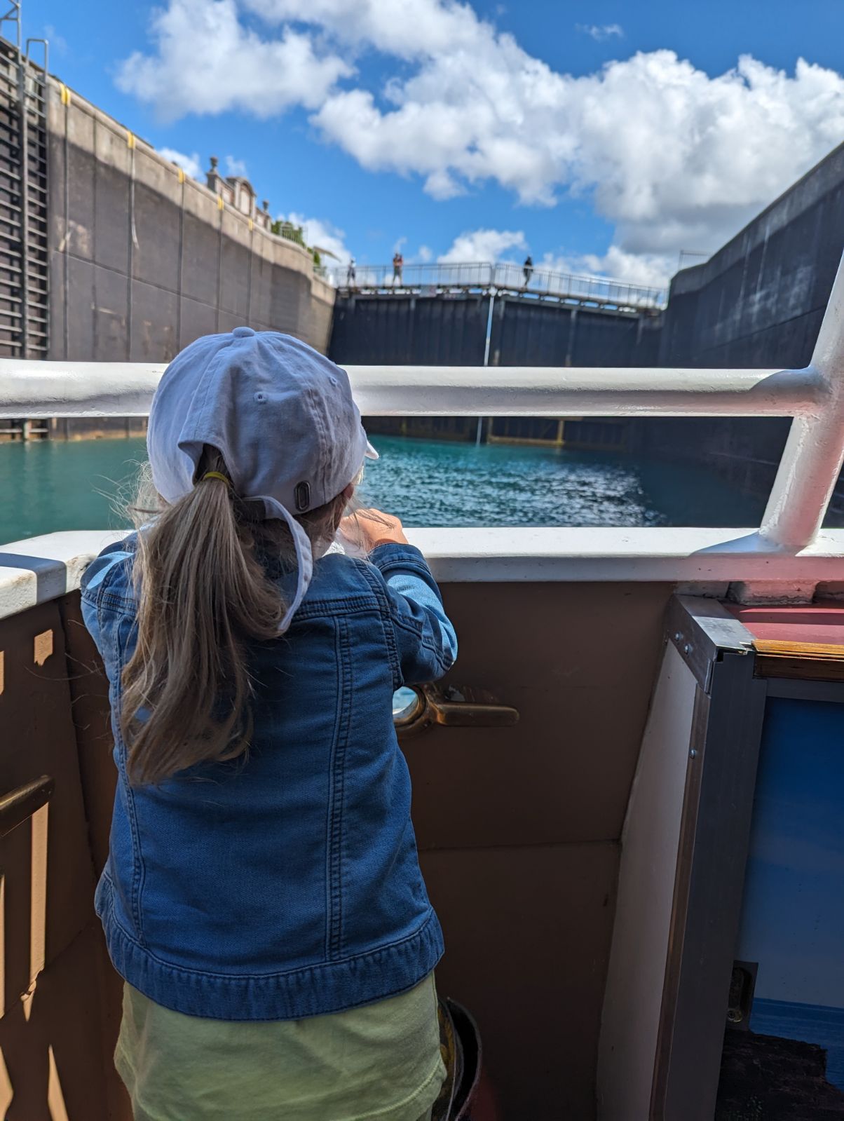 A small girl looking over the metal railing of a ship as it enters the Sault Locks, filled with shining blue-green water under a blue sky.