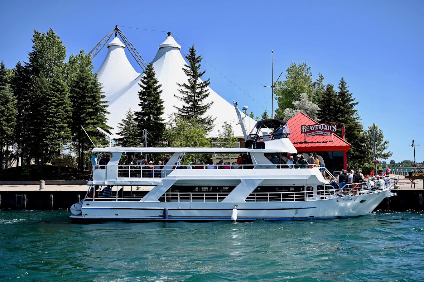 the ship "Miss Marie" docked near Roberta Bondar Park; a large white boat docked on green-blue lake water next to two tall-peaked tent pavilions that peek up above the trees. The sky is a clear, sunny blue. 