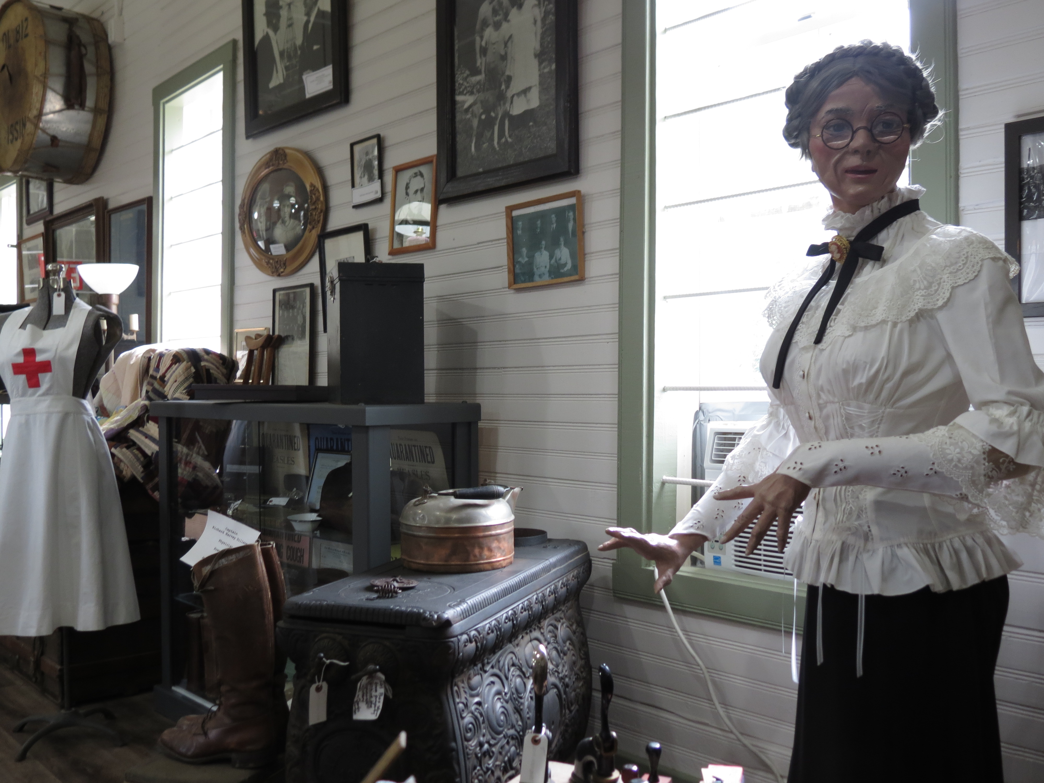 The interior of the Nipissing Township Museum; a series of artifacts on display next to tall sunlit windows, such as hanging pictures, antique clothing on mannequins, and an antique stove.