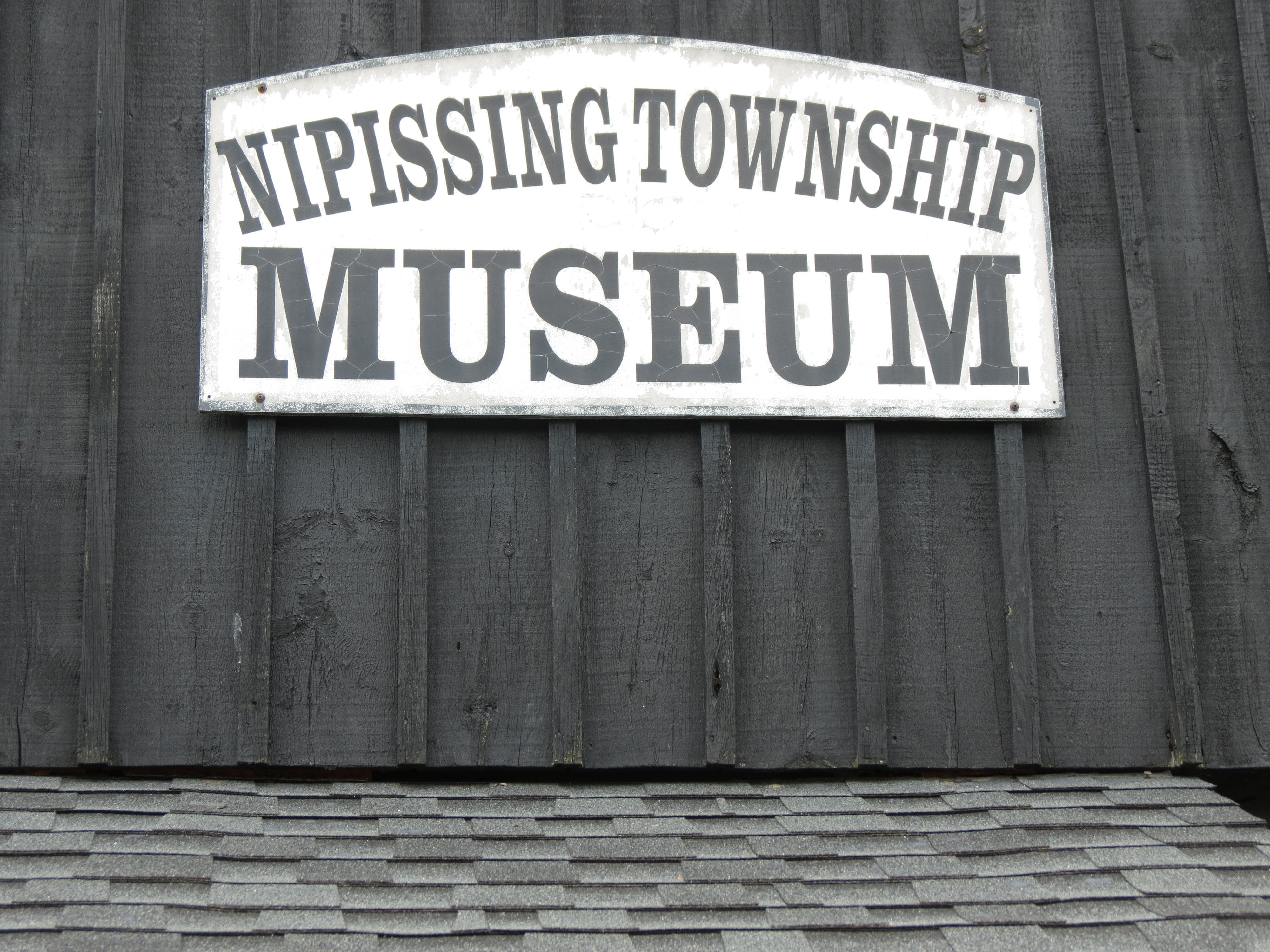 A grey and white sign above a grey shingled roof that reads "Nipissing Township Museum".