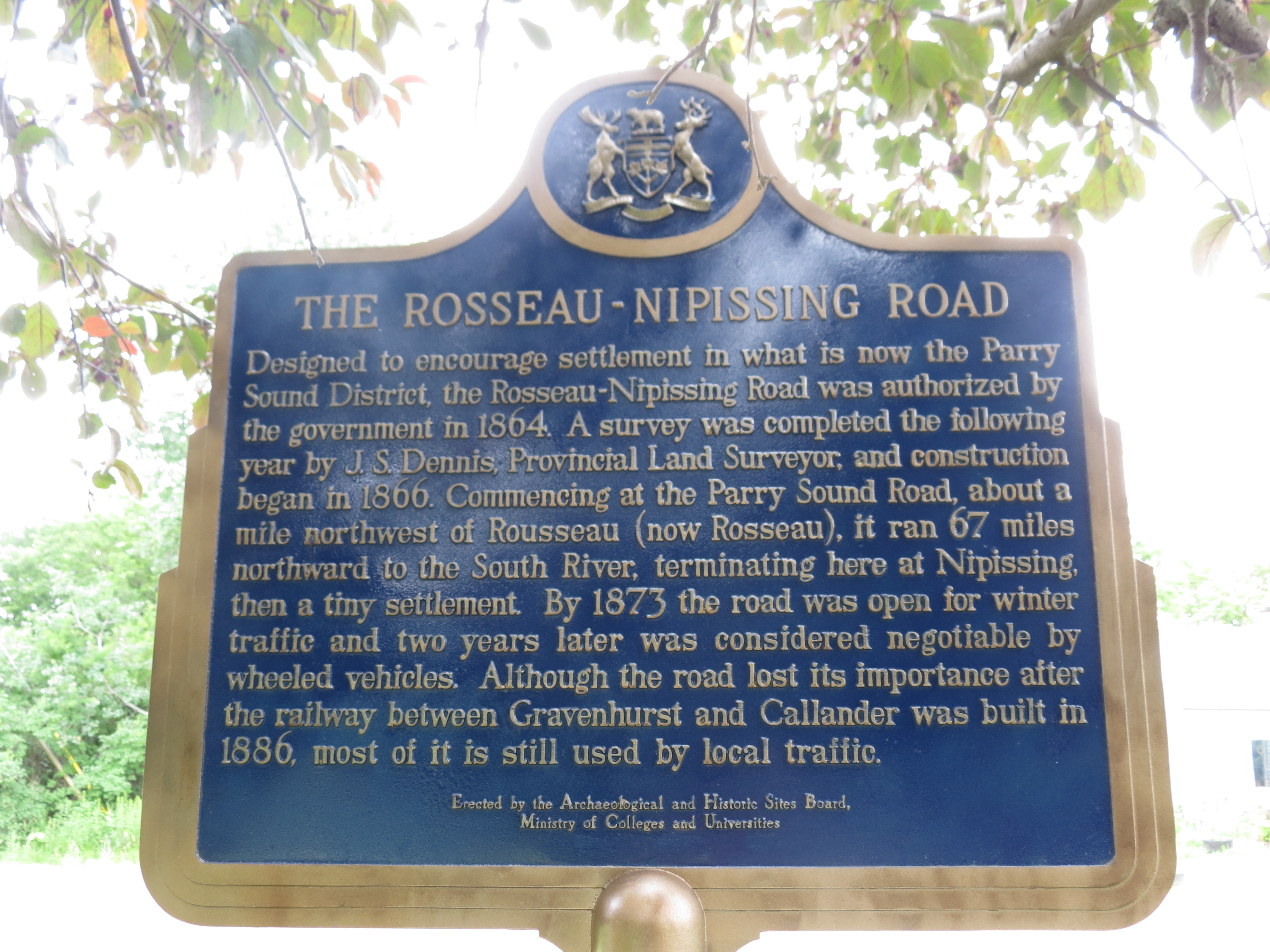 A blue and gold commemorative sign describing the authorization and construction of the Rosseau-Nipissing Road, with green foliage and sunlight in the background.