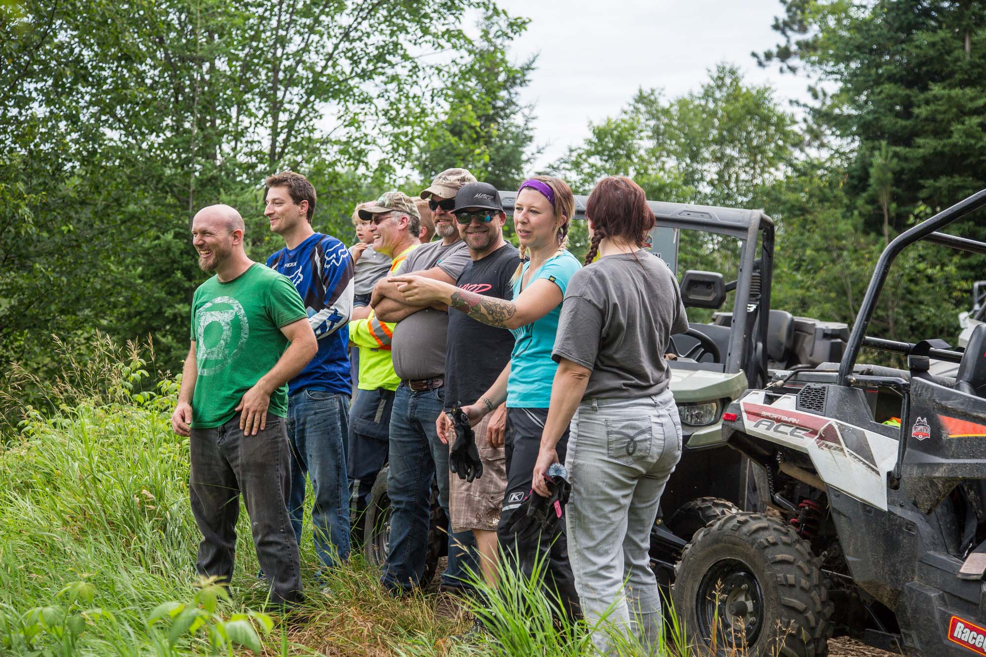 a group of smiling people laughing and talking about something out of the frame; one woman is pointing her finger as she speaks. They are standing in a forest next to their ATVs and UTVs.
