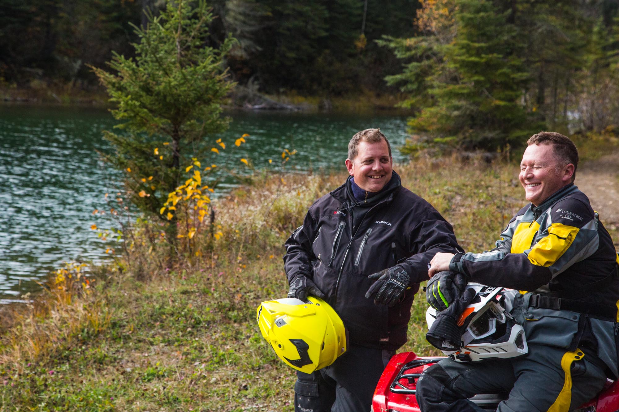 Two ATV riders talking and laughing while they are parked next to a lake surrounded by forest.
