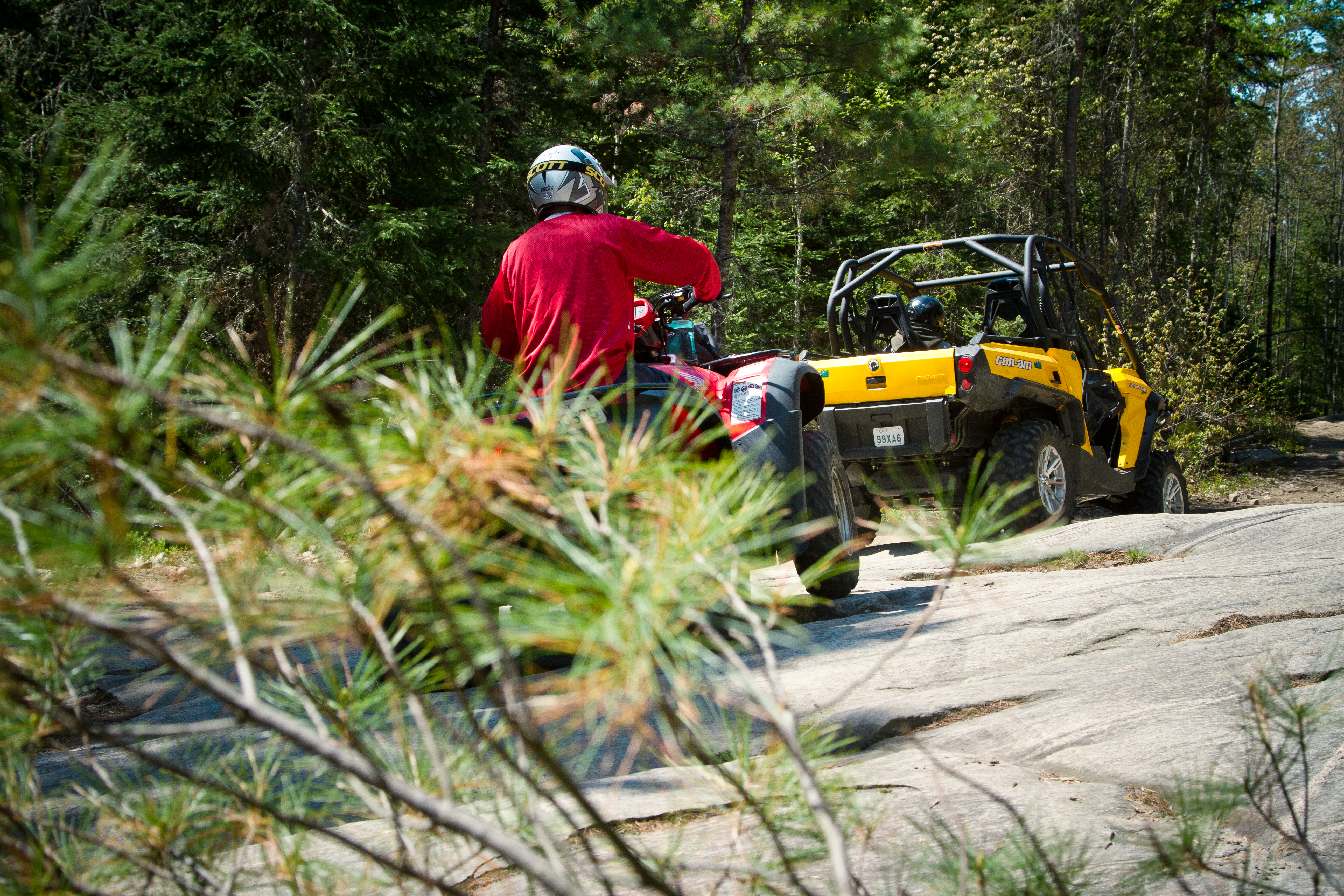 Riders driving their ATV and UTV down a smooth rock trail surrounded by pine brush and tall green forest.