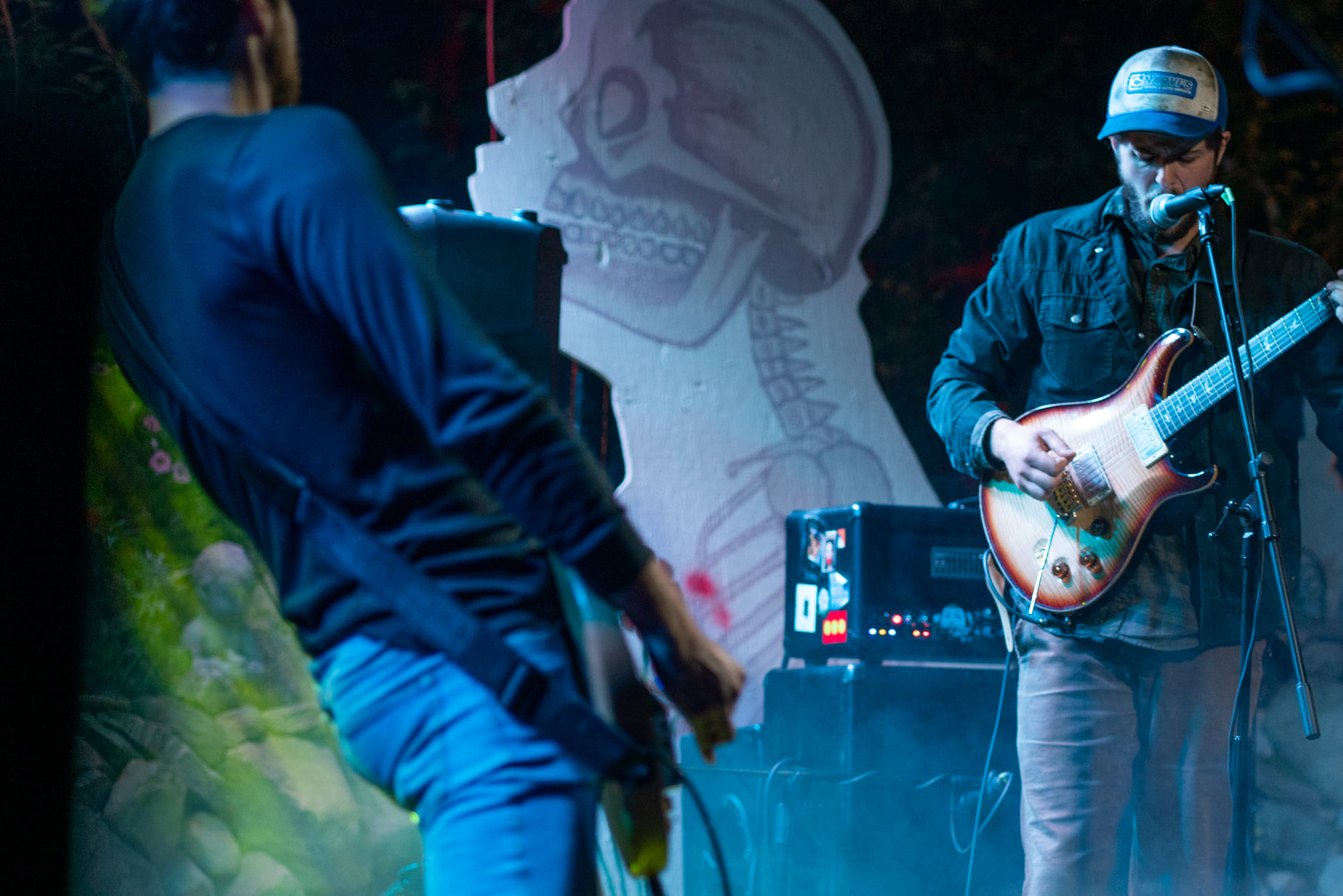 Two guitarists performing on a darkened stage with a decorative cutout of a skeleton's profile behind them.