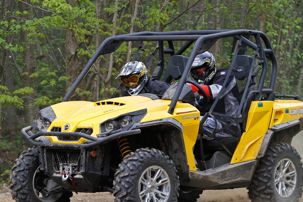 A mud-spattered UTV with two smiling, helmetted people in it driving down a forest trail.