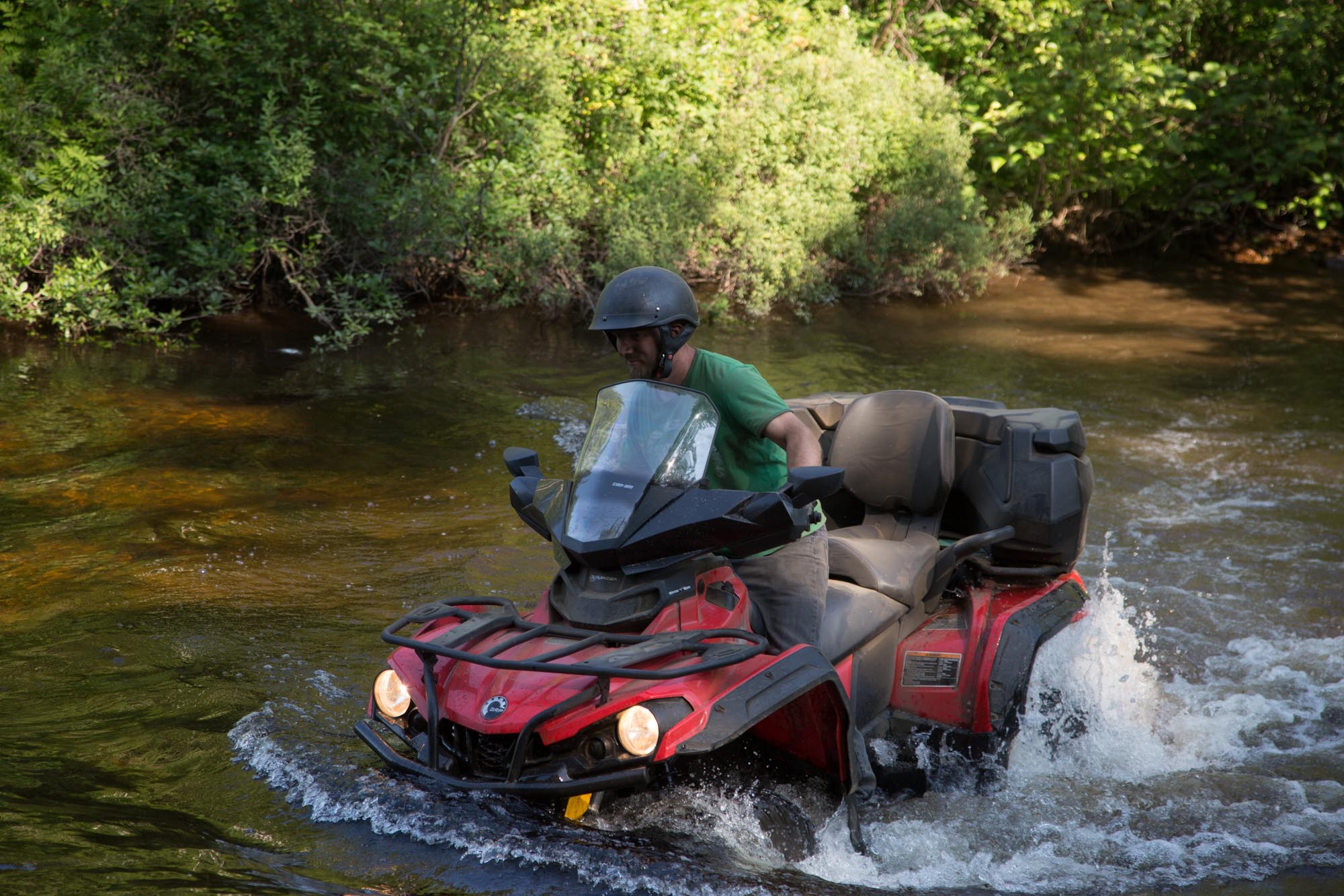 a rider drives an ATV through a stream in a dense green forest. The water is up to the tops of the tires.