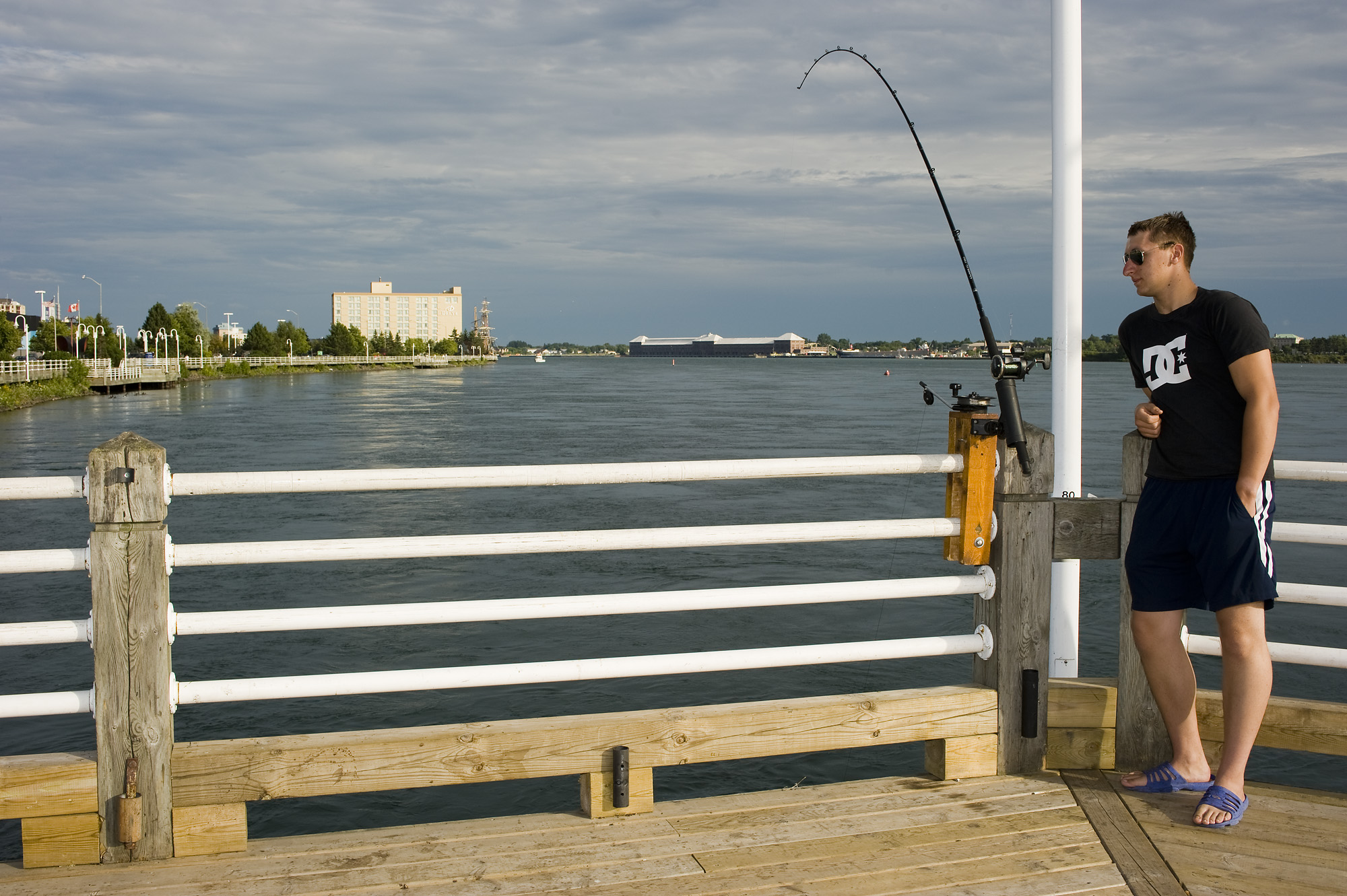 a man wearing shorts and sunglasses leans against the railing as he stands on a fishing platform in Sault Ste. Marie. He is looking at the large fishing pole that sits in a holder on the railing, the pole bending slightly with the weight on the line.