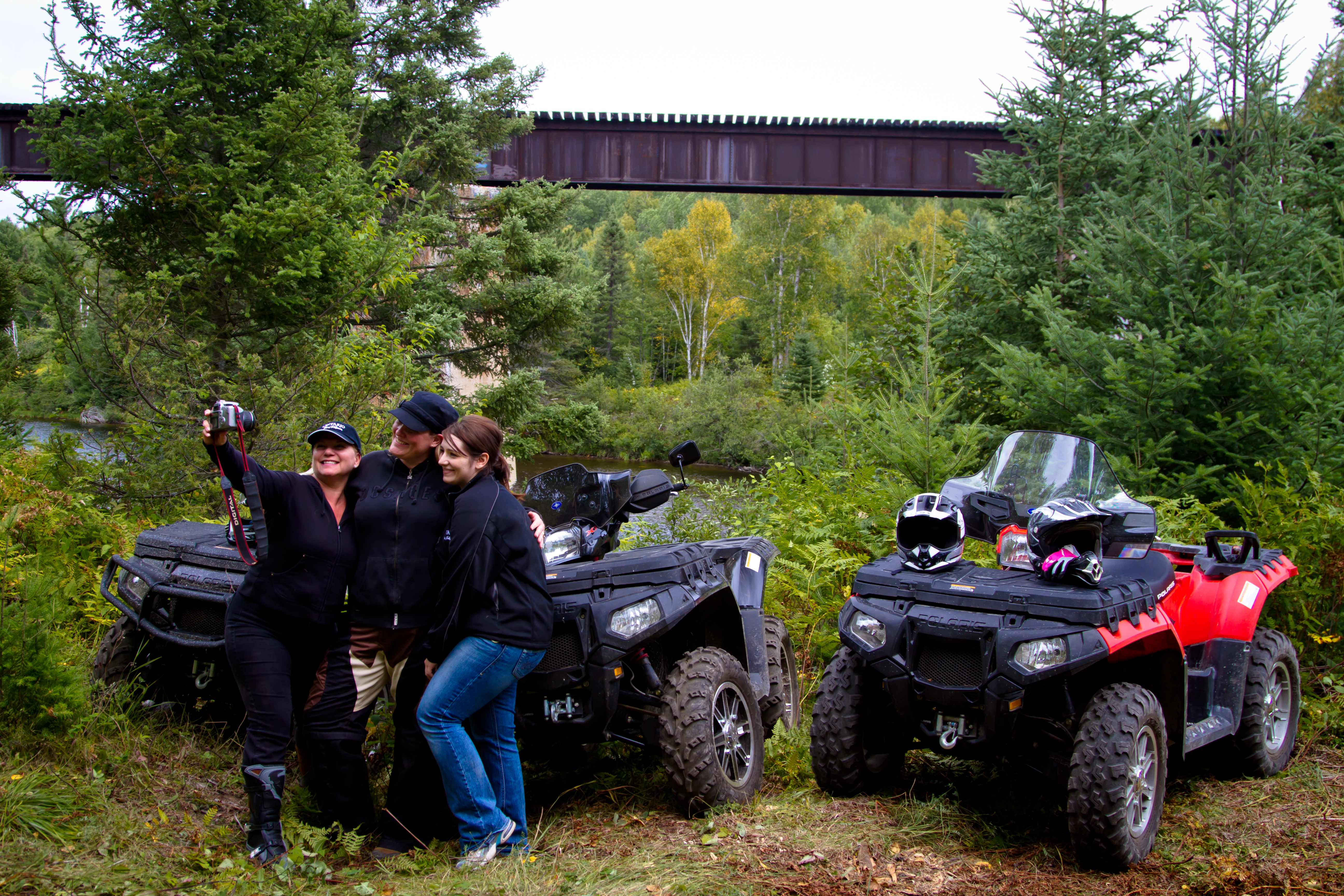 3 friends smiling and hugging together while one holds a camera at arm's length to shoot a photo. Their 3 ATVs and green forest and field are behind them. 