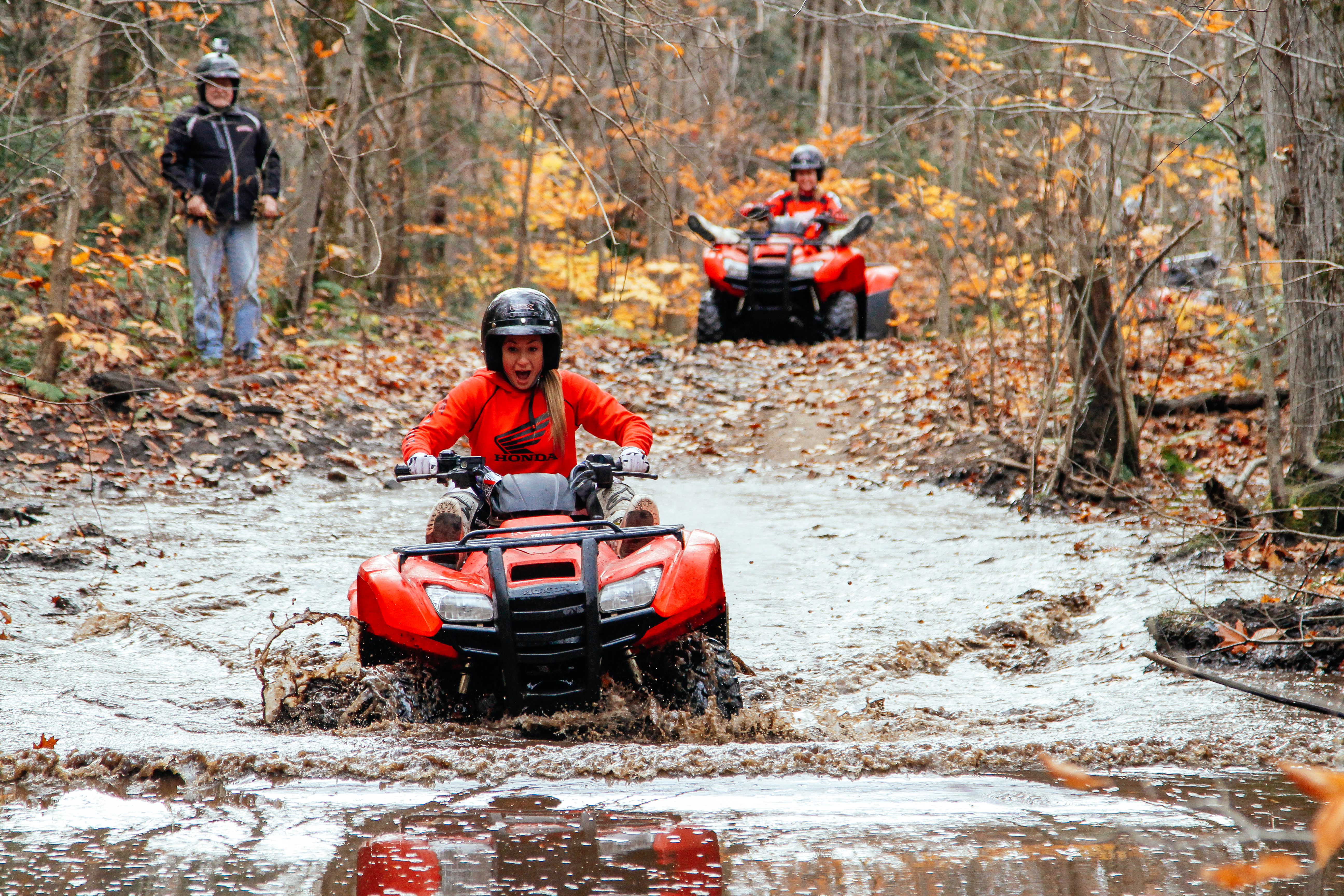 A woman riding an ATV with a shocked look on her face as she splashes through a mud hole in the middle of a forest trail on a grey autumn day. The muddy water is up to the top of the ATV's tires. 