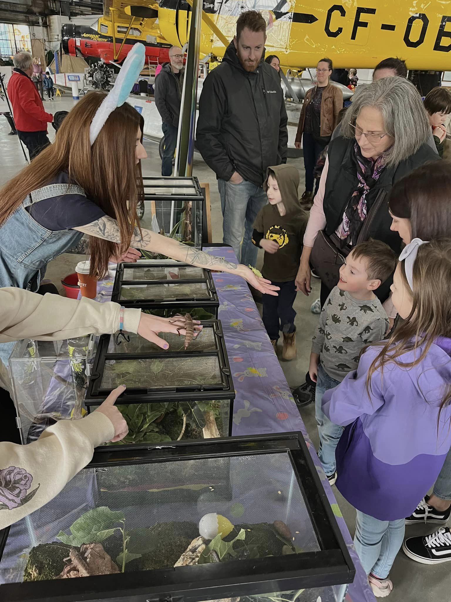 Children looking at an insect being held out for them by a red head in bunny ears.