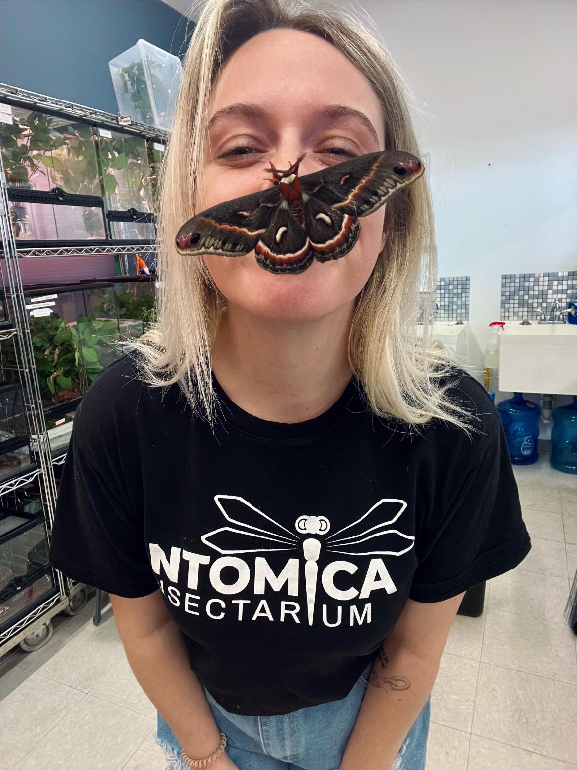 A white blonde woman with a large moth on her nose. Her black t-shirt says Entomica Insectarium.