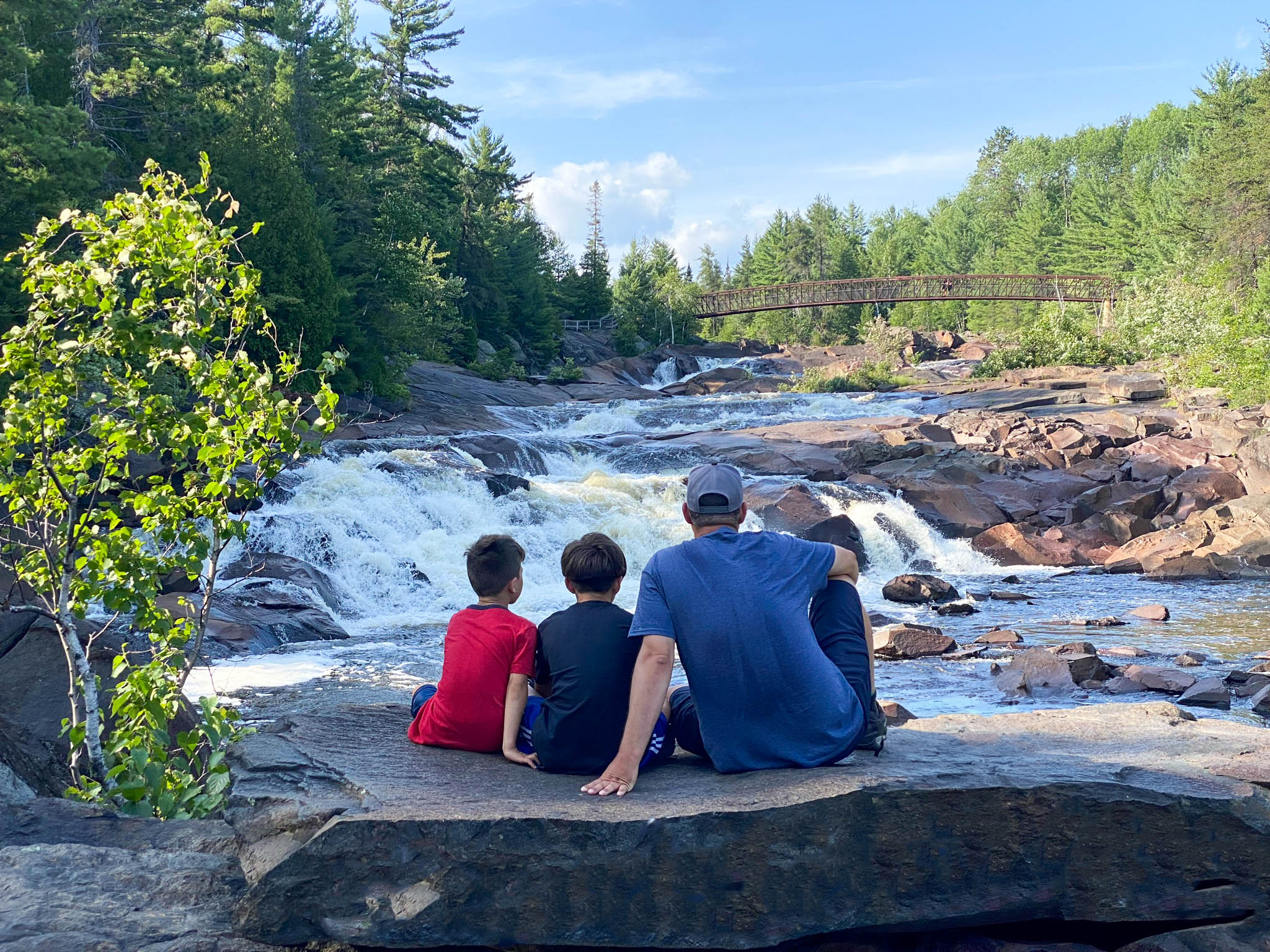A father and two young sons sitting on a large rock together, looking at High Falls on a sunny summer day.