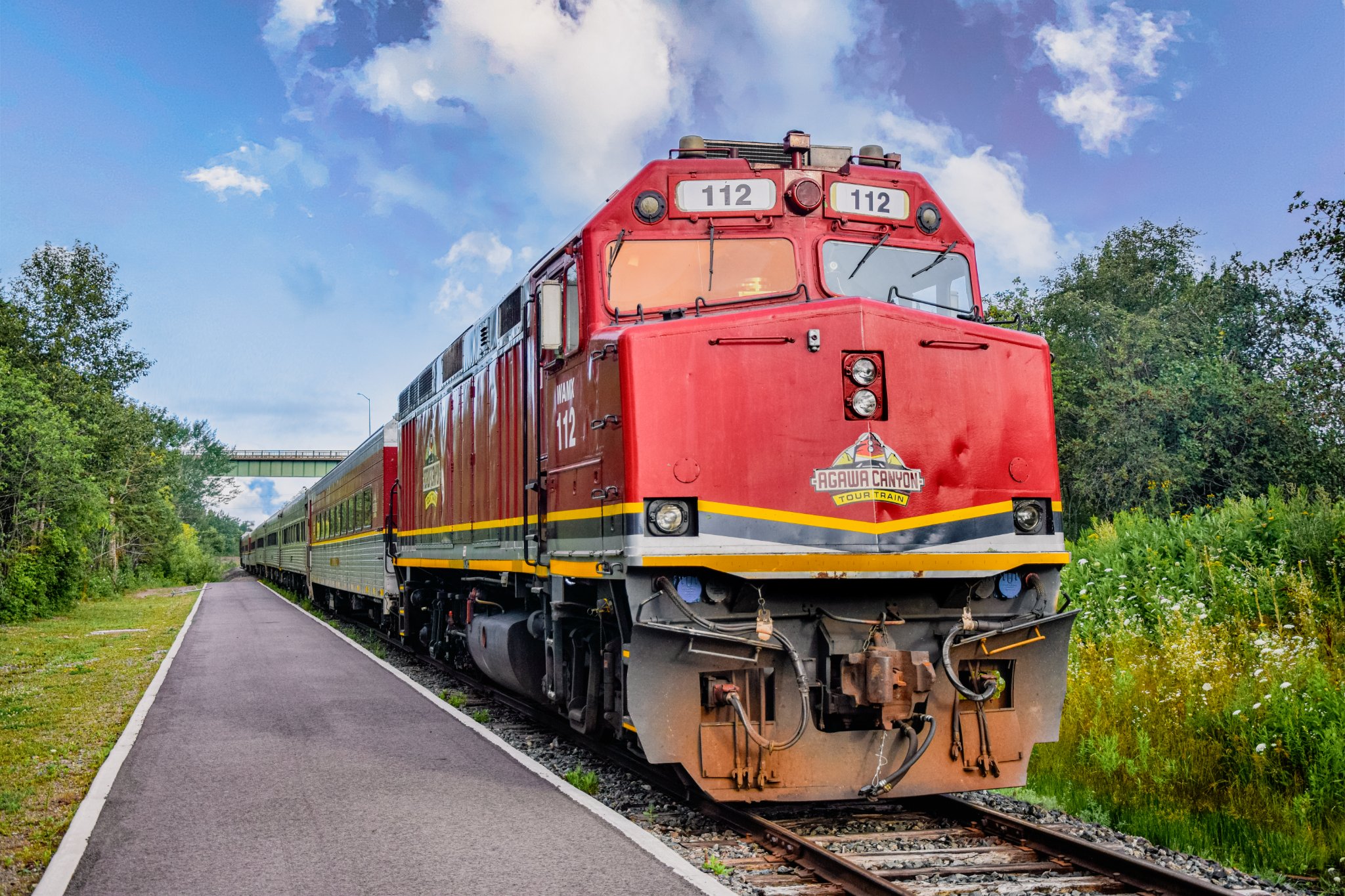 A red passenger train gliding down the track through lush green forested countryside under a bright blue sky.