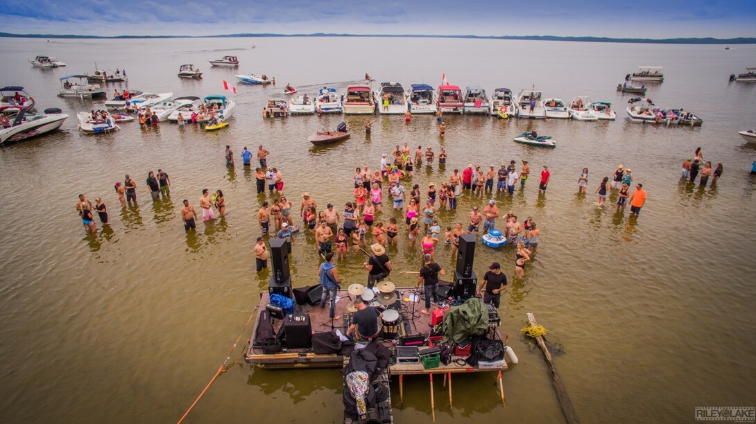 a crowd of people standing in shallow water on a sandbar in front of a stage for the Berrio at the Sandbanks concert. Boats are parked in the water all around.