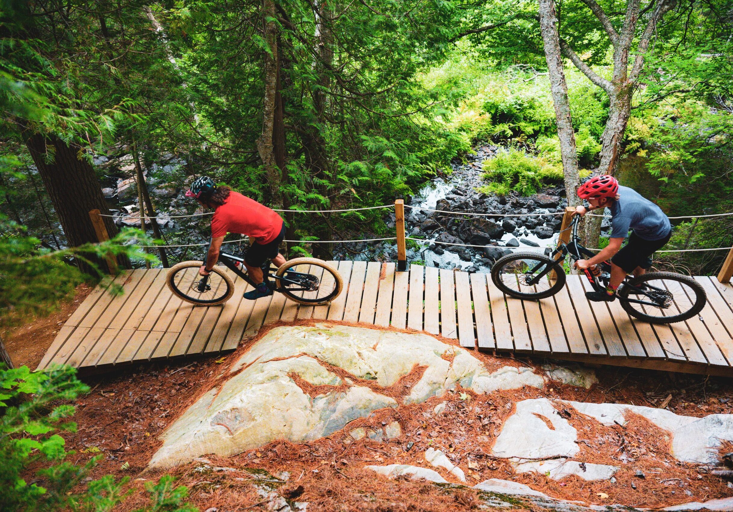 Cyclists head across a narrow wooden bridge on a gravel trail surrounded by large smooth boulders and lush, dense green forest.