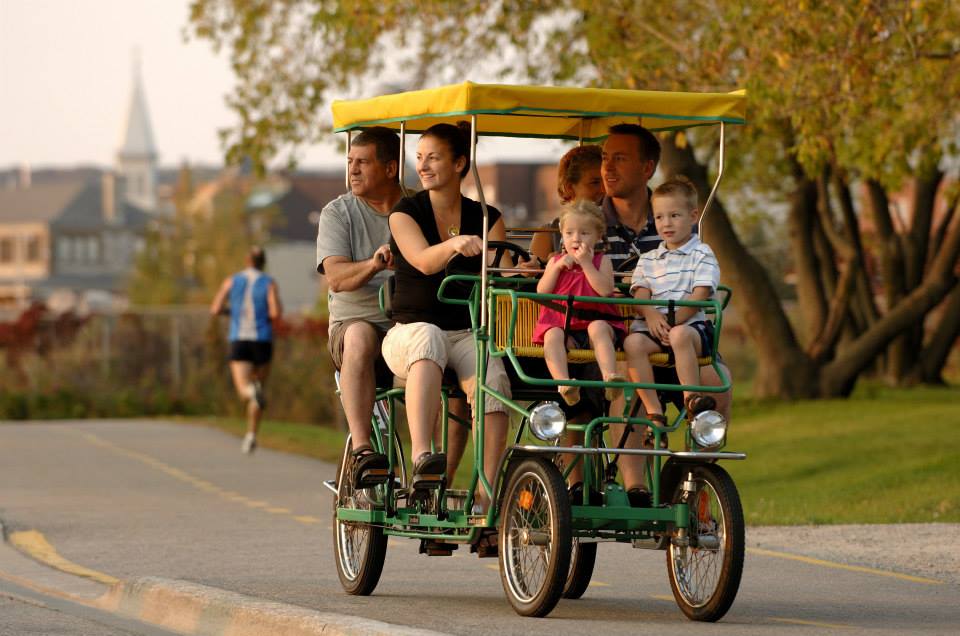 a smiling family riding together in a 4-wheeled multi-person pedal cart along the North Bay waterfront in the late afternoon.