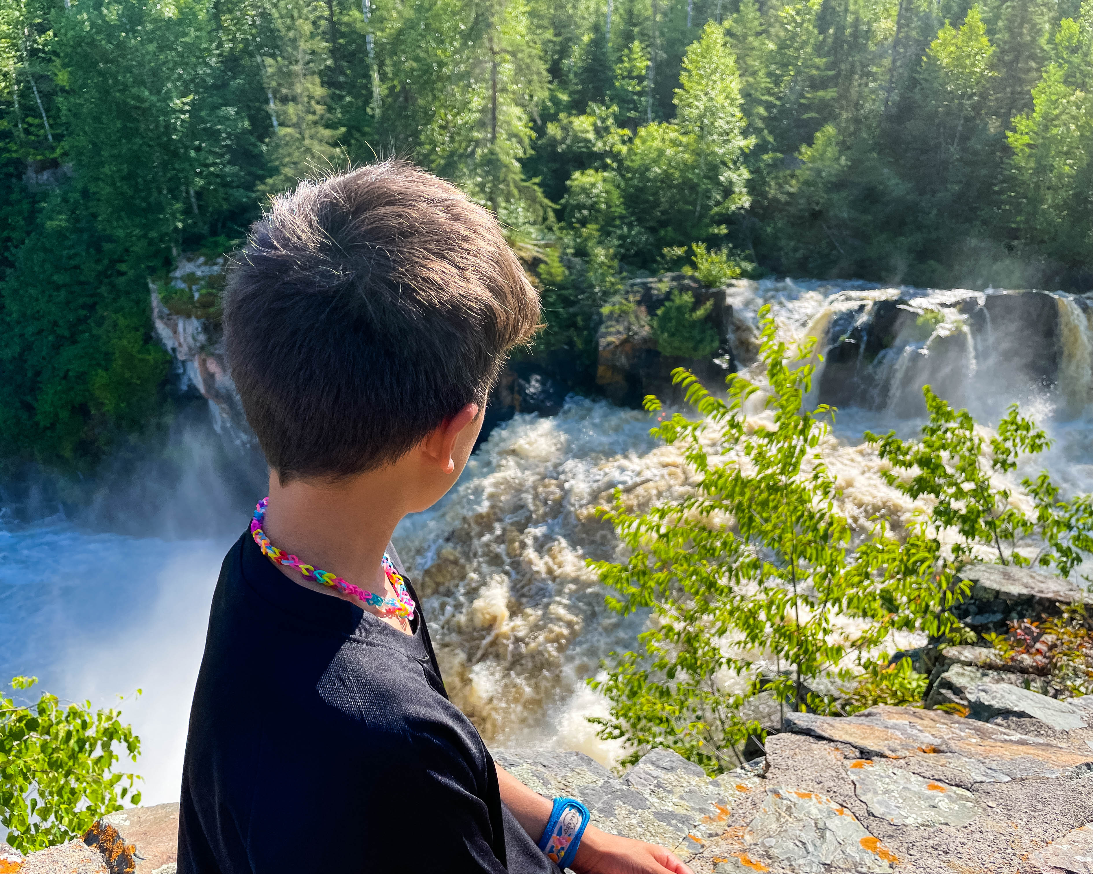 A boy stands at a high point on a rocky cliff, looking at the falls at Kap Kig Iwan Provincial Park on a summer day. 