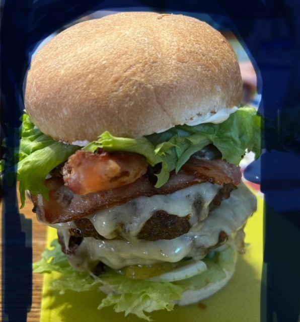 A close-up of the Mattagami Rivers Fries signature burger which is called the River Burger. It looks like it has 2 patties layered with cheese on top of lettuce, pickles and onions and topped with bacon, lettuce and their signature sauce.