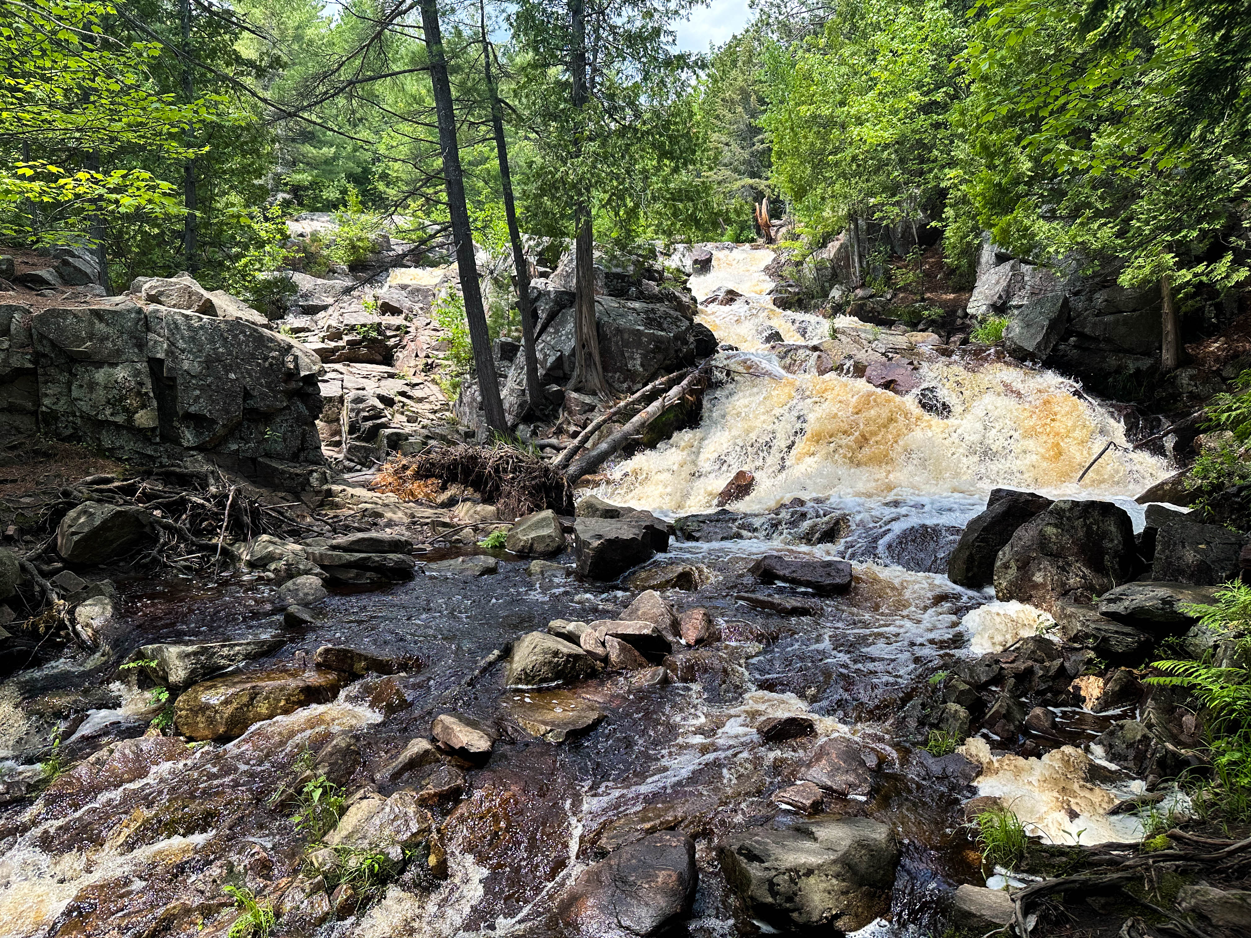 Duchesnay Falls; a low, bubbling waterfall along a bouldered riverbed in a thick green forest.