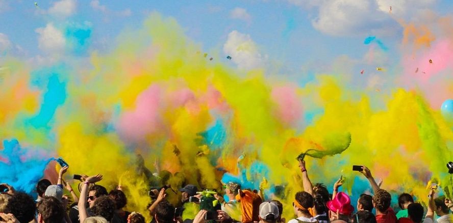 a crowd of people waving their hands above their heads as they throw a rainbow of multicoloured clouds of powdered pigments into the air at Holi Fest.