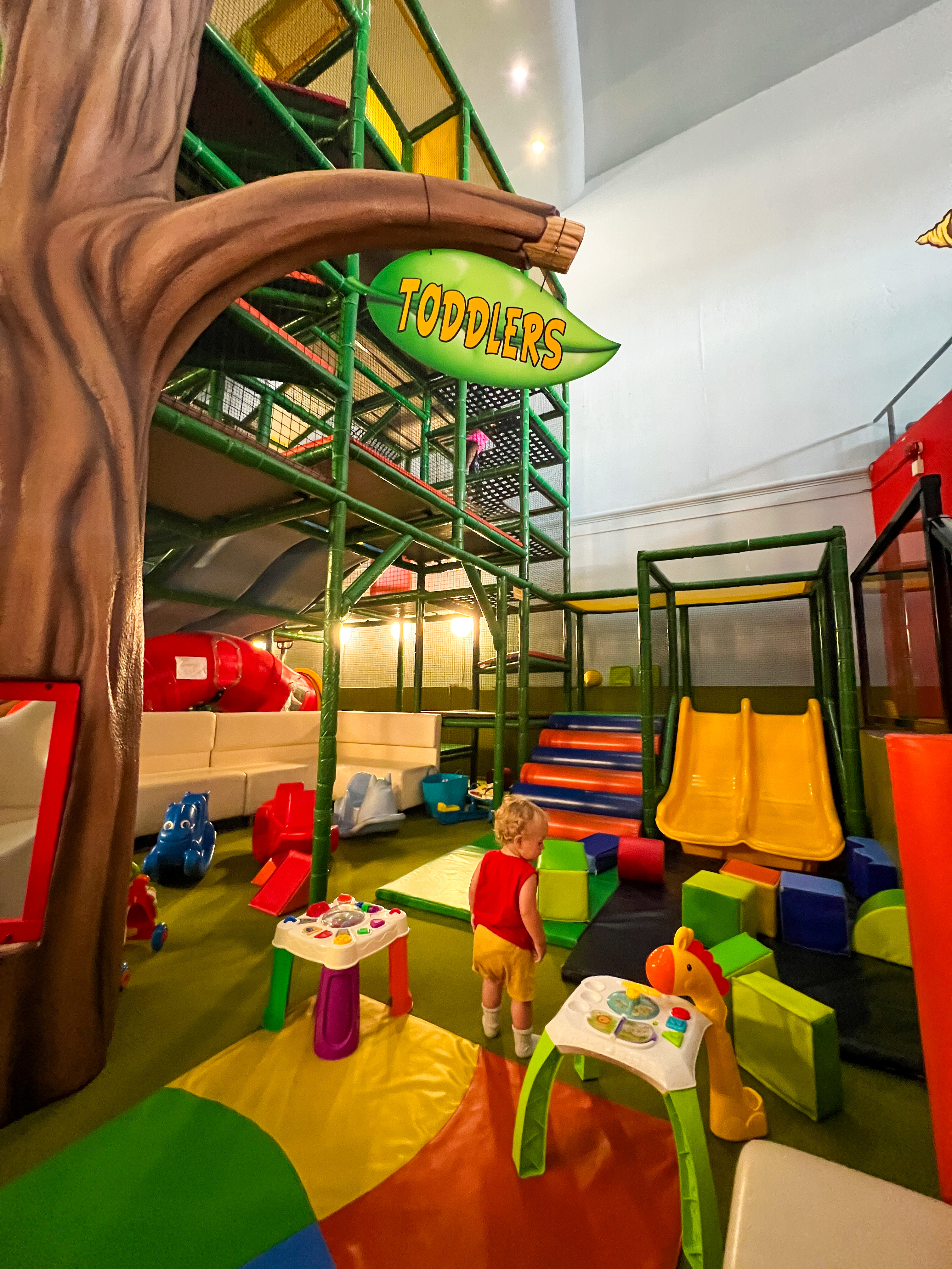 A colourful indoor playground with a climbing apparatus and a toddler in a red shirt.