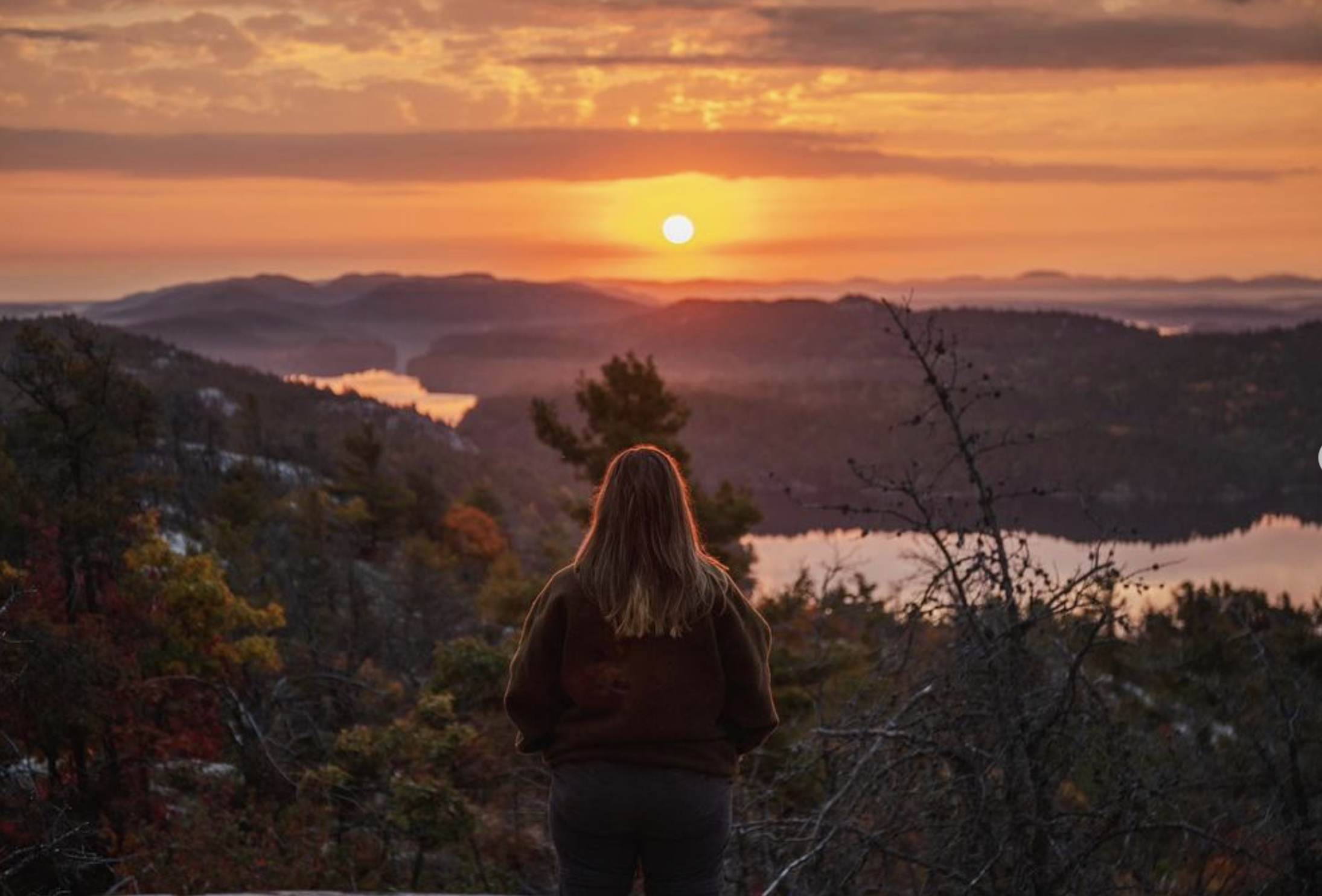 La Cloche; a woman looking out over an orange sunrise over a rocky, forested valley with a large lake.