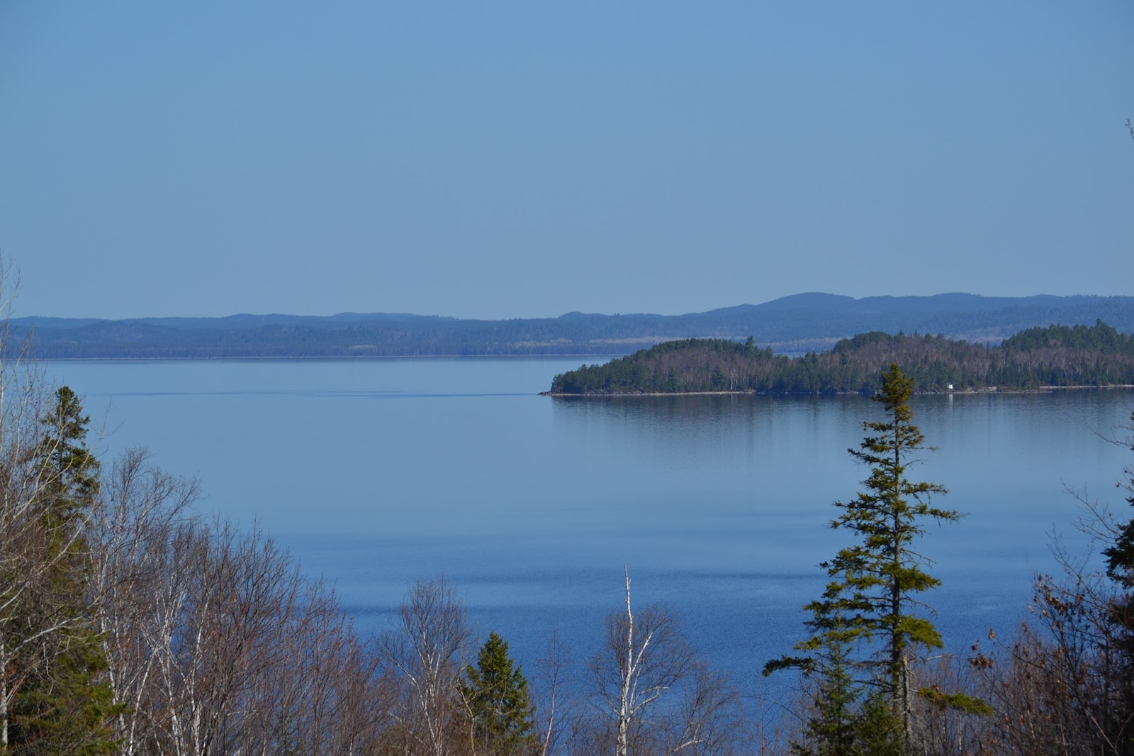 Lake Wanapitei; a very large, still, blue lake under a misty blue sky, surrounded by forest and with a low, sloping and rocky far bank on the horizon.